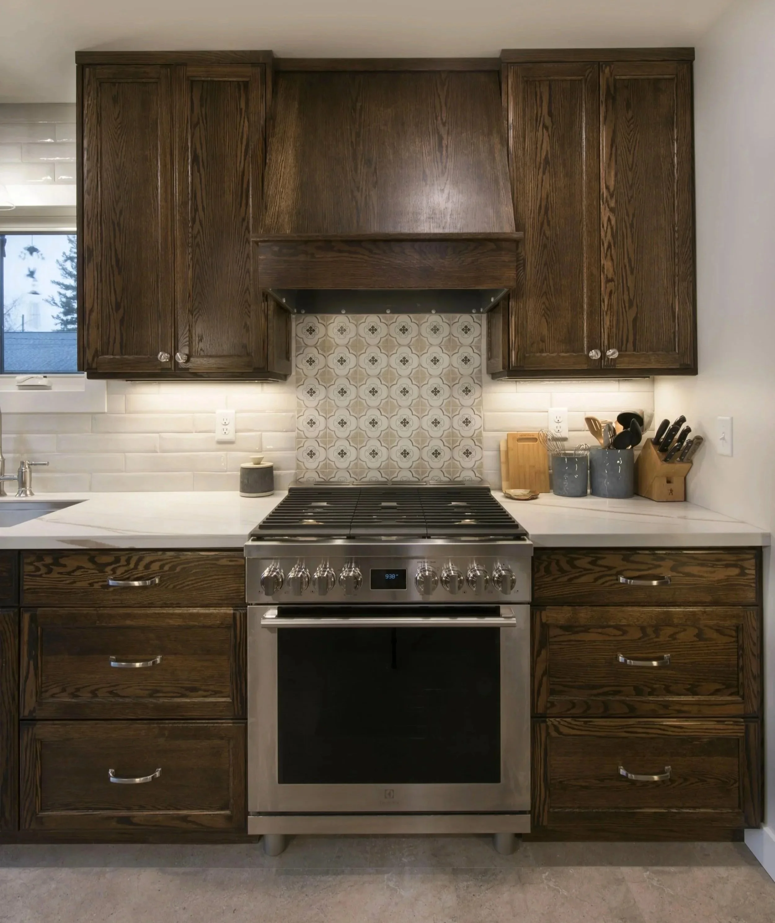 Kitchen with dark wood cabinetry, stainless steel stove, white countertop, beige and patterned tile backsplash, window with trees outside, and kitchen utensils on the counter.