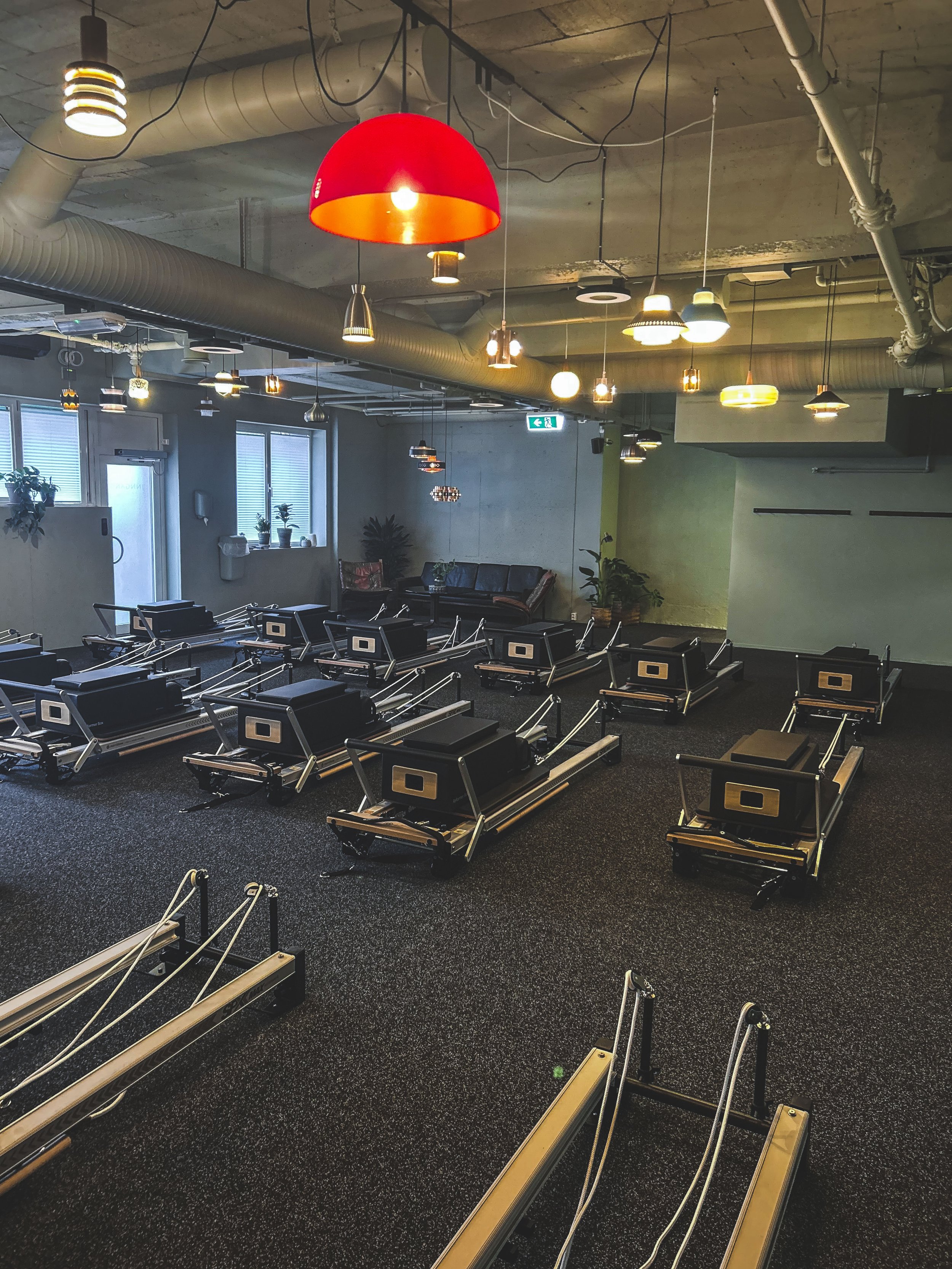 Rom med treningsutstyr, flere pilates- eller treningsbenker, stoler, planter og lyse dekorative lamper i taket.