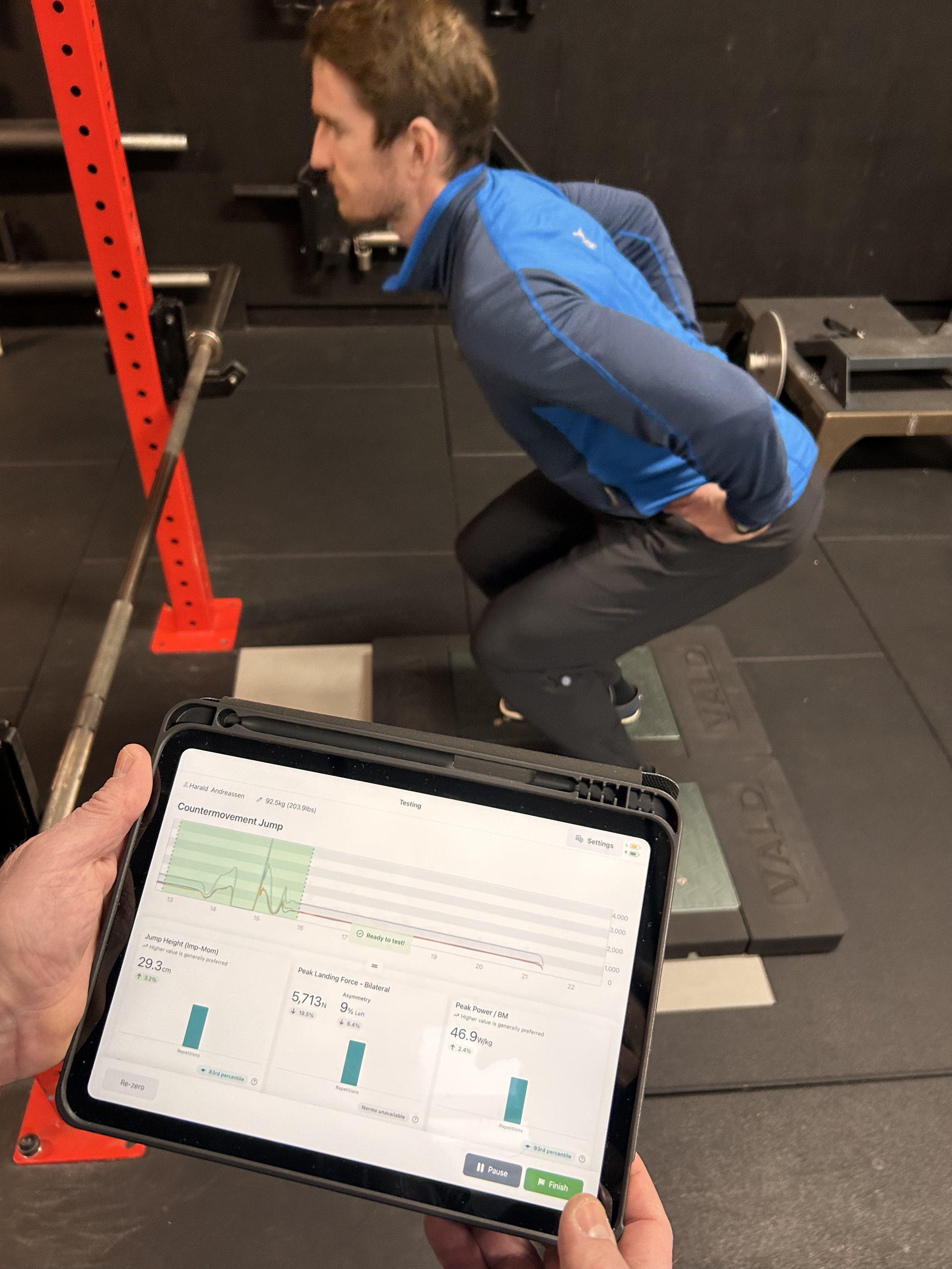 En mann gjør en knebøy på treningsplattform i treningsstudio, mens en annen person holder et nettbrett med en treningsanalyse på skjermen.