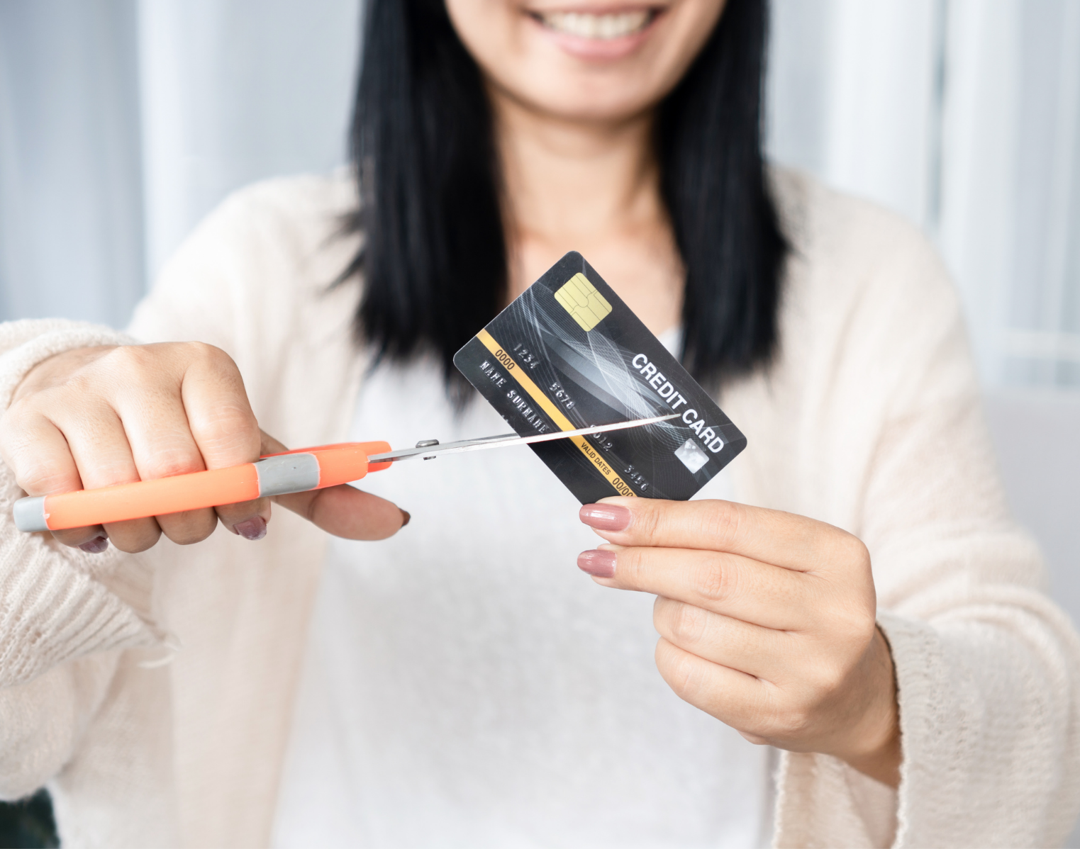 A woman with black hair smiling while holding a credit card with her left hand and a large orange scissors with her right hand, cutting the credit card.