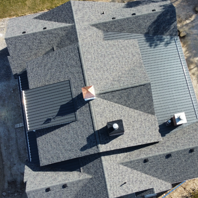 Aerial view of a house roof with multiple sections, different vent pipes, and roofing materials, including shingle and metal panels. An aerial view of a custom home with 24 gauge standing seam metal roof