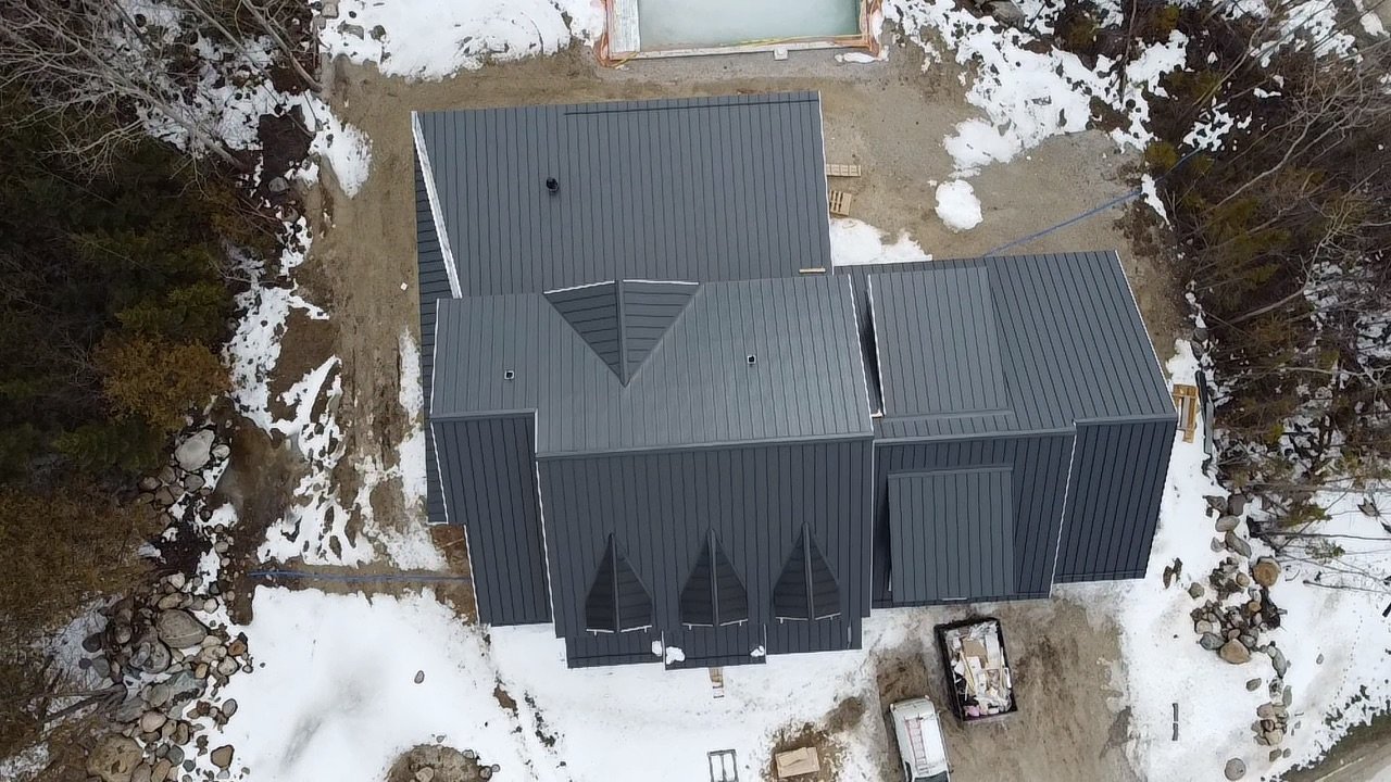 An aerial view of a house with a gray metal roof, surrounded by snow-covered ground and trees.