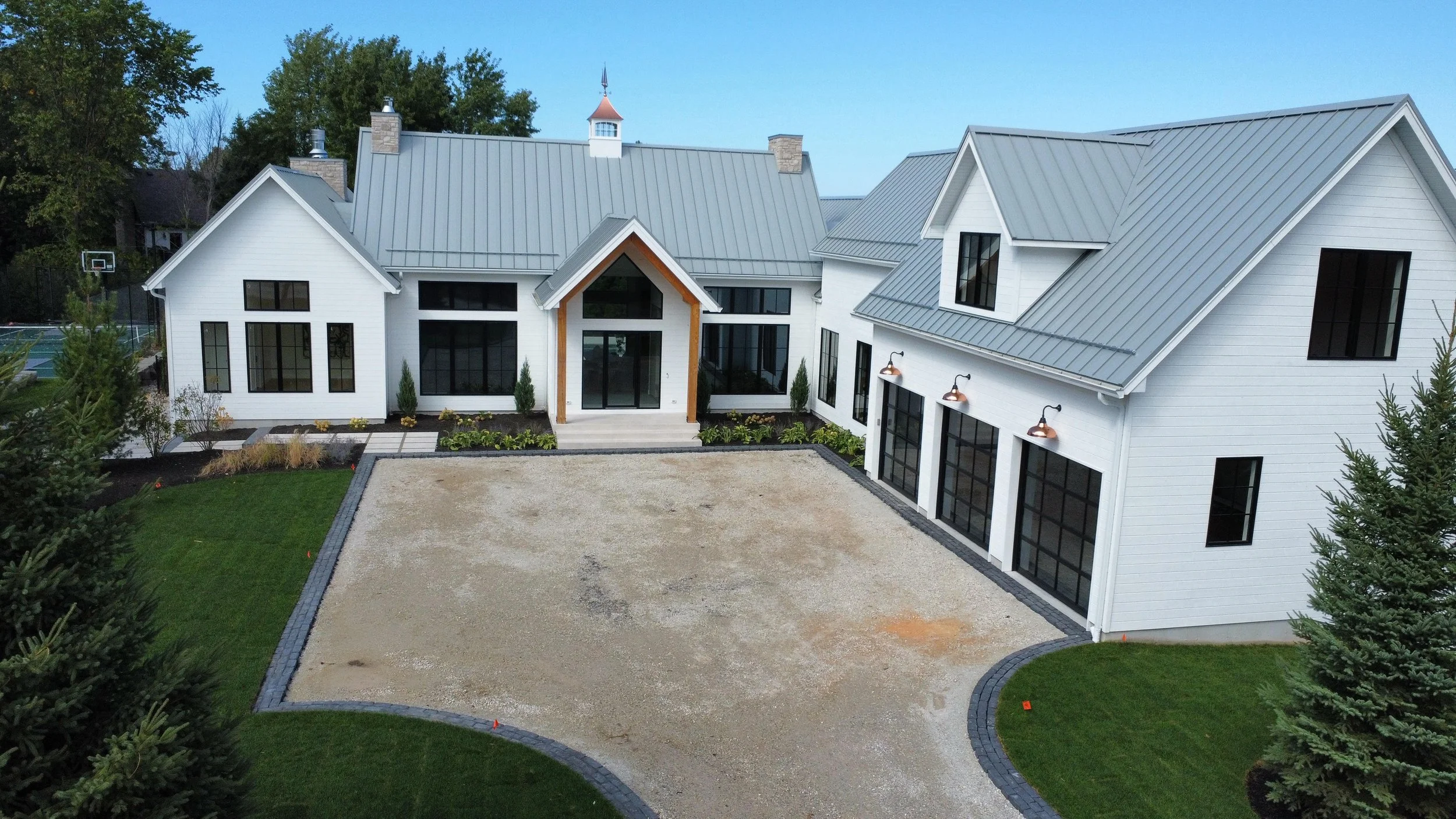 An aerial view of a custom home with 24 gauge standing seam metal roof, A large, white modern house with a gray metal roof, black window frames, and a landscaped front yard with a concrete driveway.