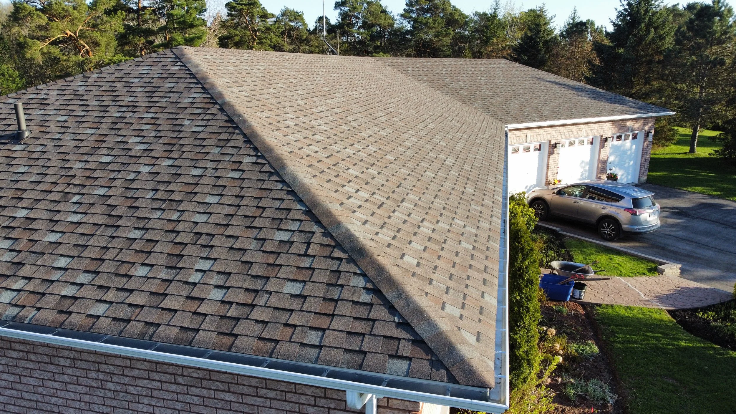 An aerial view of a house with a brown shingle roof, a garage with three white doors, a parked silver SUV, and a landscaped lawn with trees in the background.