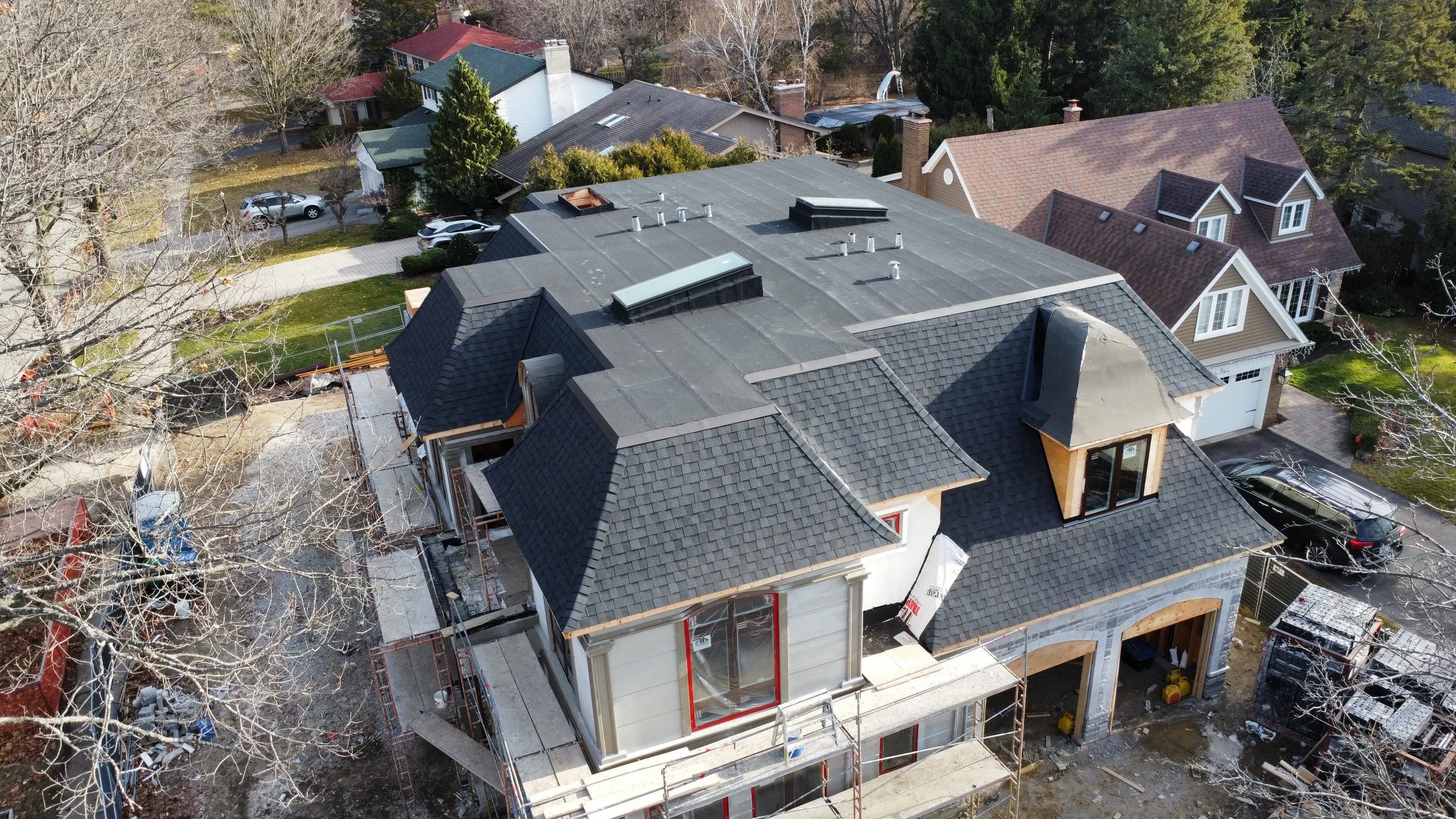 Aerial view of a house under construction with a black shingle roof, surrounded by neighboring houses and trees, some leafless, and construction materials and scaffolding around the building.