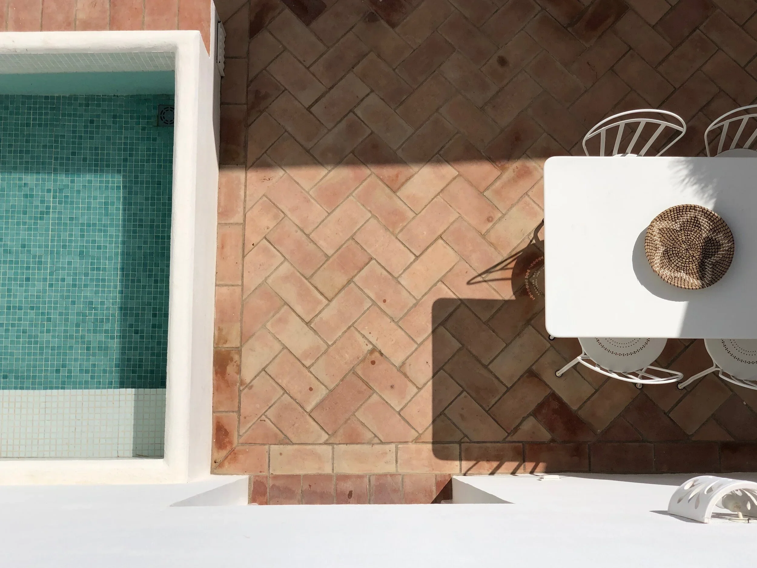 Imagem de uma área externa com piso de tijolos, uma piscina com revestimento de azulejos verdes, uma mesa branca com uma tigela de palitos de comida e quatro cadeiras brancas ao redor da mesa.