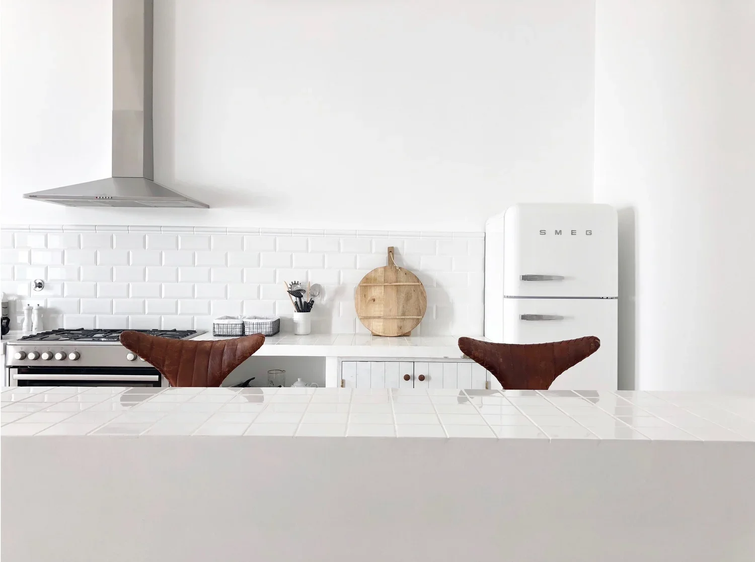 Cozinha branca com duas cadeiras de couro marrom, uma geladeira SMEG branca, um fogão de metal, uma parede de azulejos brancos e utensílios de cozinha