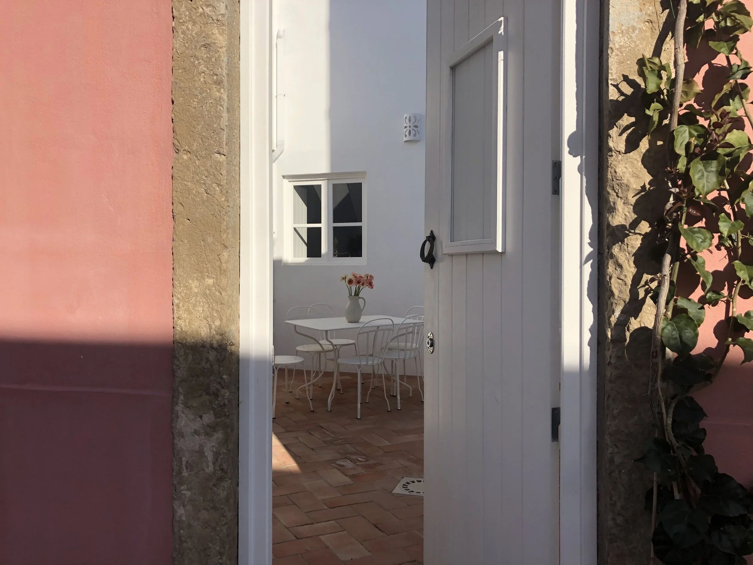 Visão de uma varanda com uma mesa branca, quatro cadeiras brancas e um vaso com flores rosas, vista através de uma porta branca aberta. Paredes de fundo em branco e janela também branca.