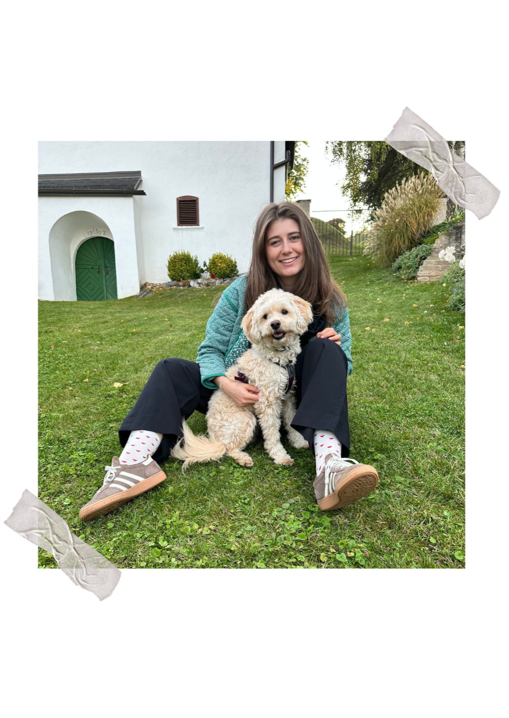 Hebamme Apollonia sitzt lächelnd im Garten mit einem kleinen, cremefarbenen Hund namens Lotte vor ihr, im Hintergrund eine weiße Hauswand mit grüner Tür und Gartenpflanzen.