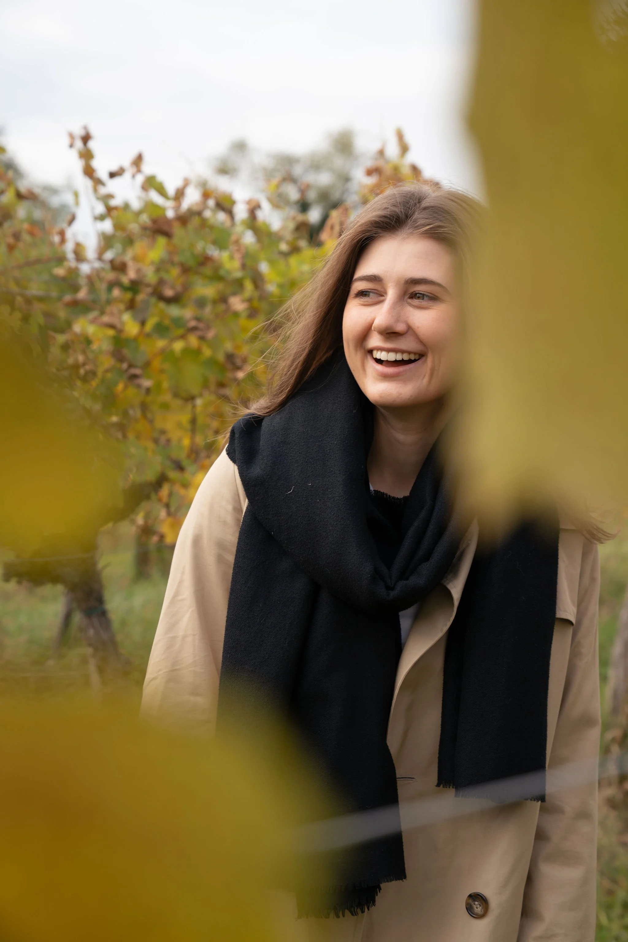 Hebamme Polla trägt einen beigen Mantel und einen schwarzen Schal, steht in einem Weingarten in Ruppersthal im Herbst, umgeben von blattverfallenen Weinreben.