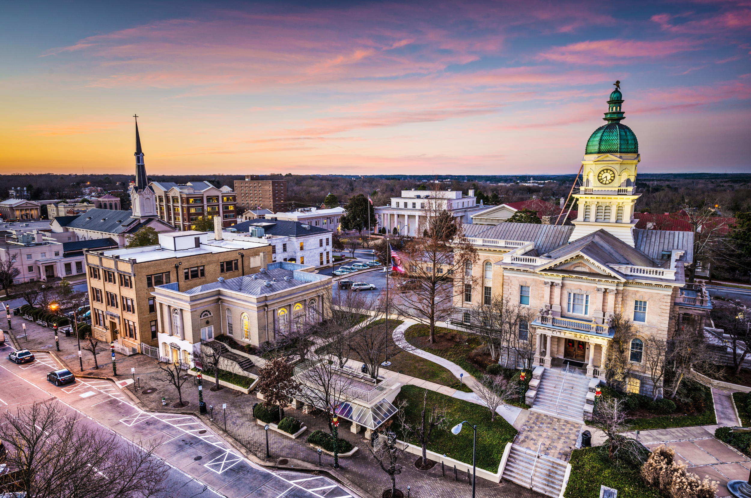 Aerial view of Athens Georgia at sunset, featuring a prominent courthouse with a clock tower, a park with a pathway, and surrounding buildings, some with pointed roofs and a small parking lot in foreground.