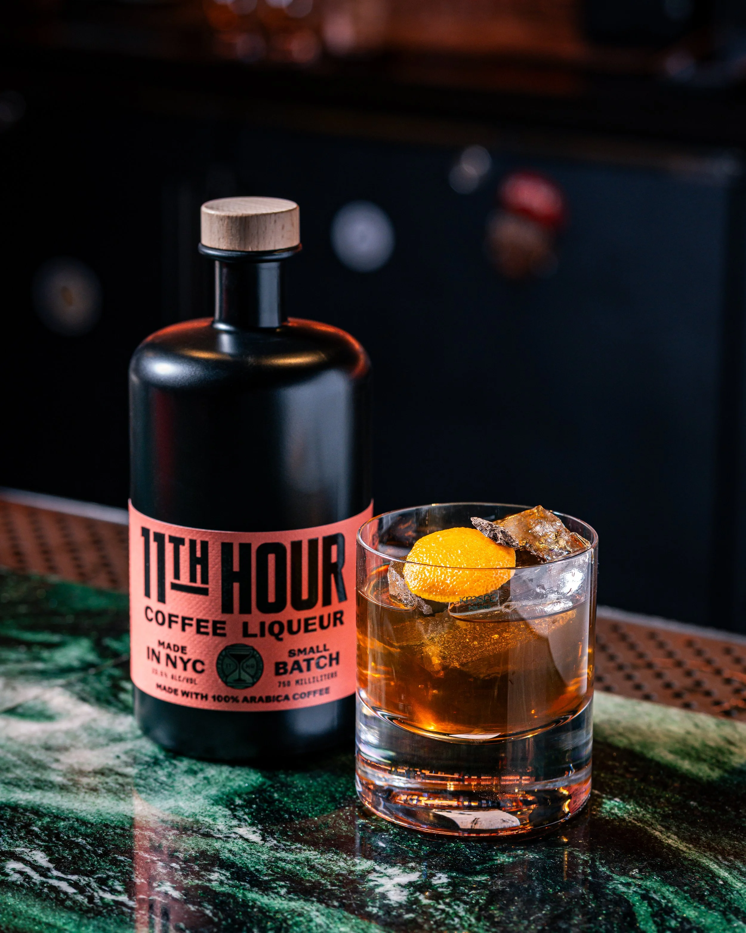 A bottle of 11th Hour Coffee Liqueur and a glass filled with a dark cocktail, ice cubes, and a lemon twist on top, on a marble countertop.