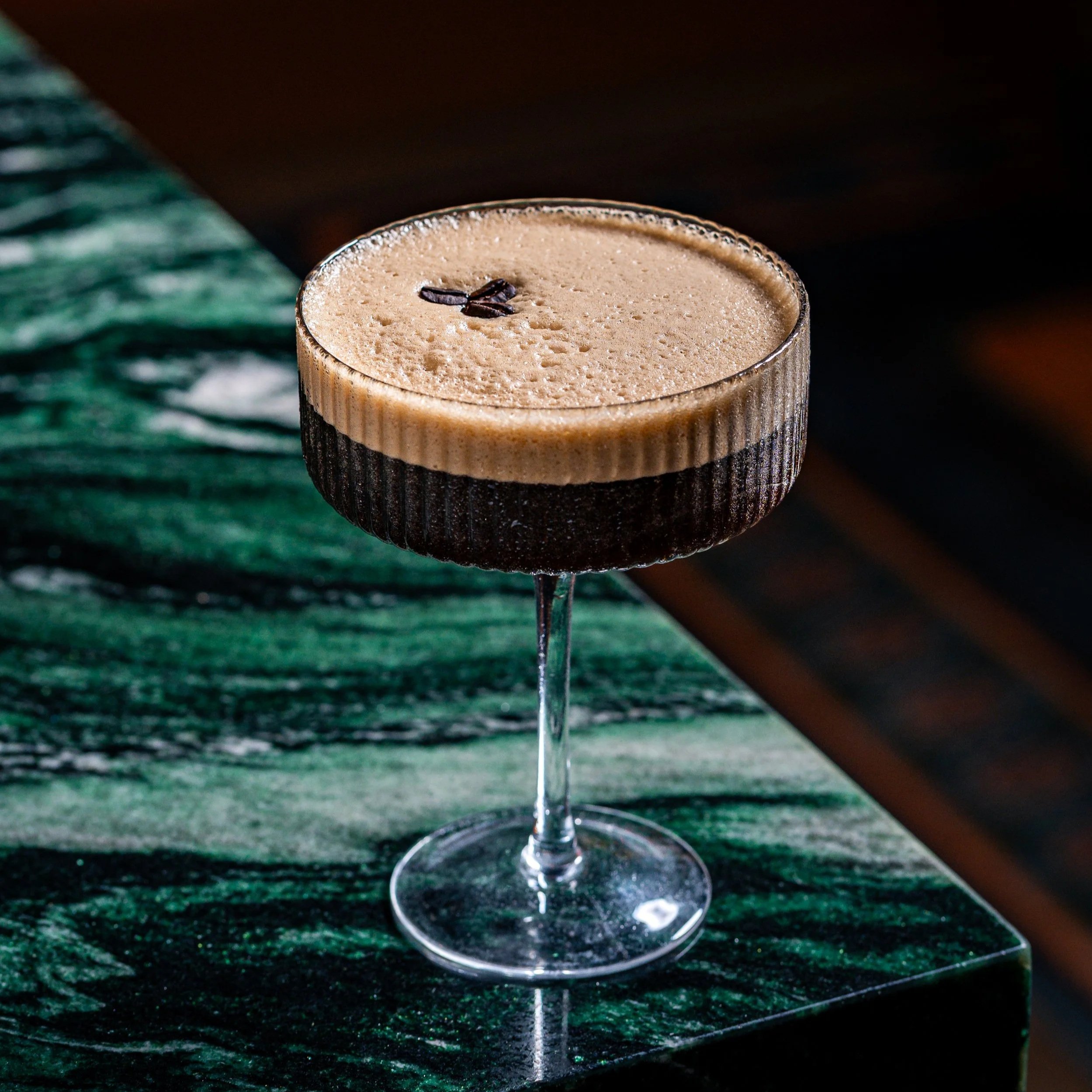 Chocolate coffee drink with foam topping and a small coffee bean garnish in a stemmed glass on a dark green marble surface.