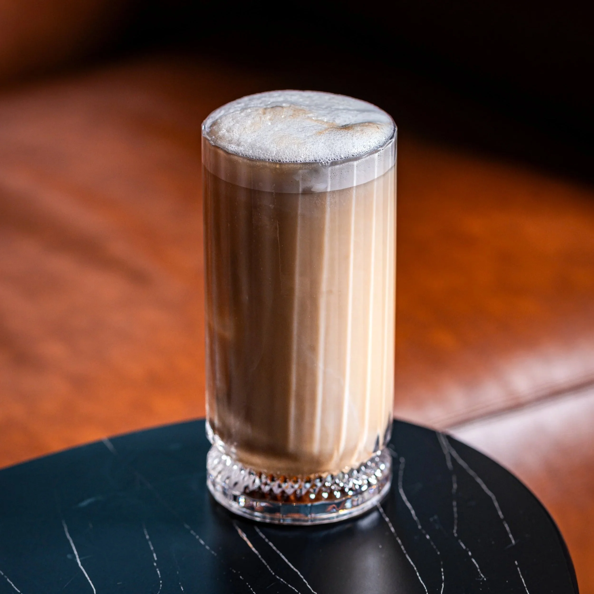 A tall glass of iced coffee with foam on top placed on a dark table.