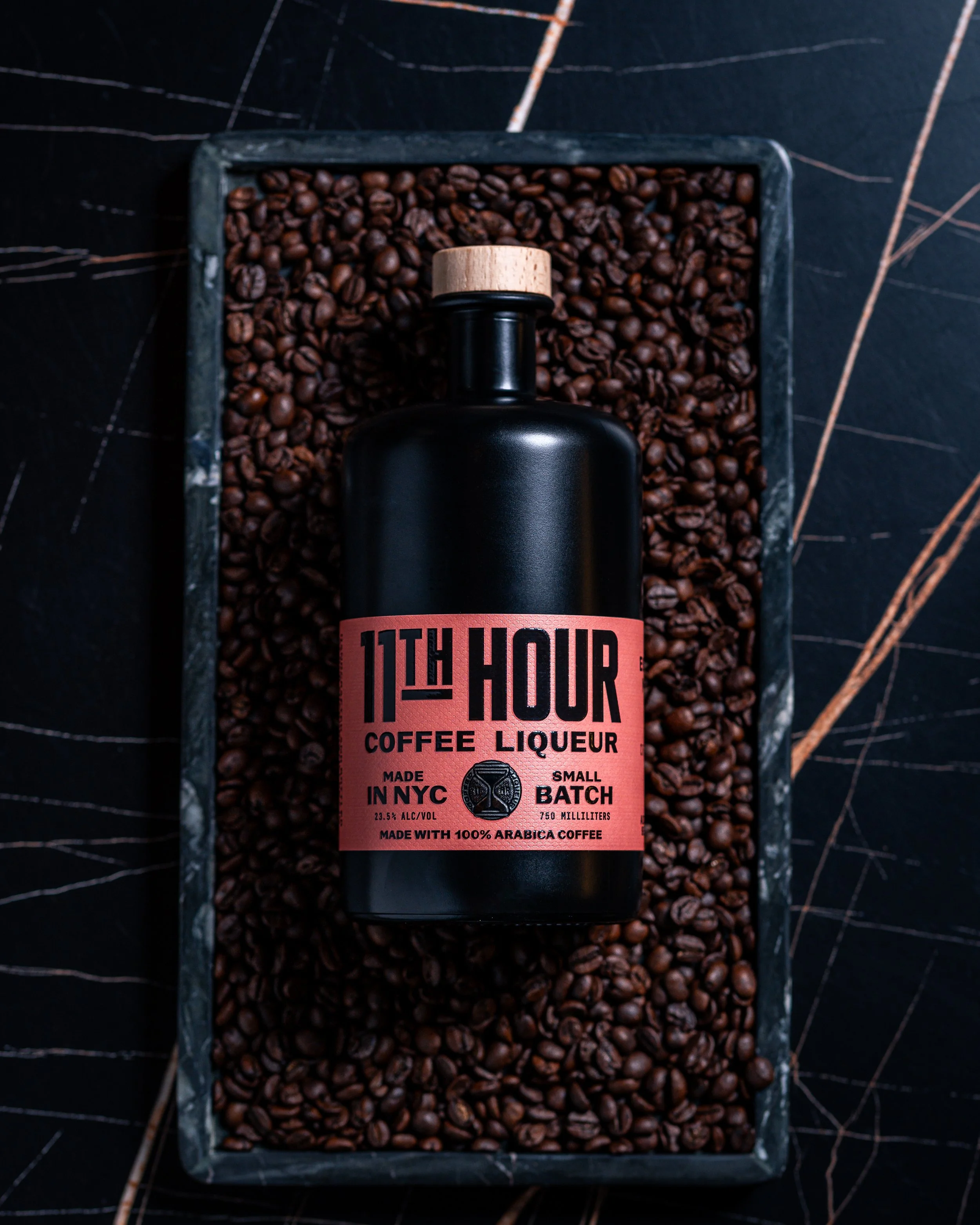 Black bottle of 11th Hour coffee liqueur with a pink label, placed on a tray filled with coffee beans.