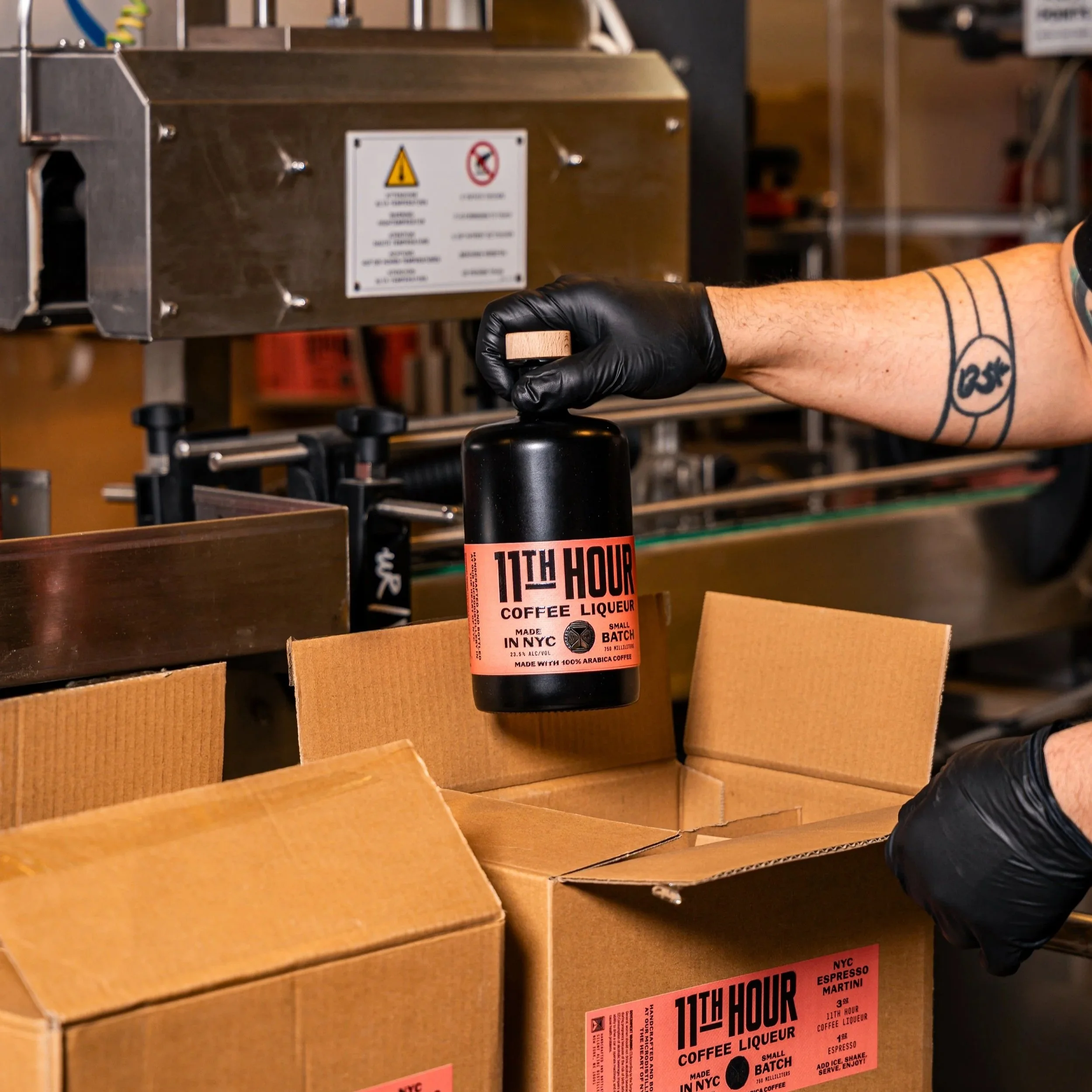 Person with tattooed arm wearing a black glove holds a black bottle labeled "11TH HOUR COFFEE LIQUEUR" over open cardboard boxes in a warehouse or packaging area.