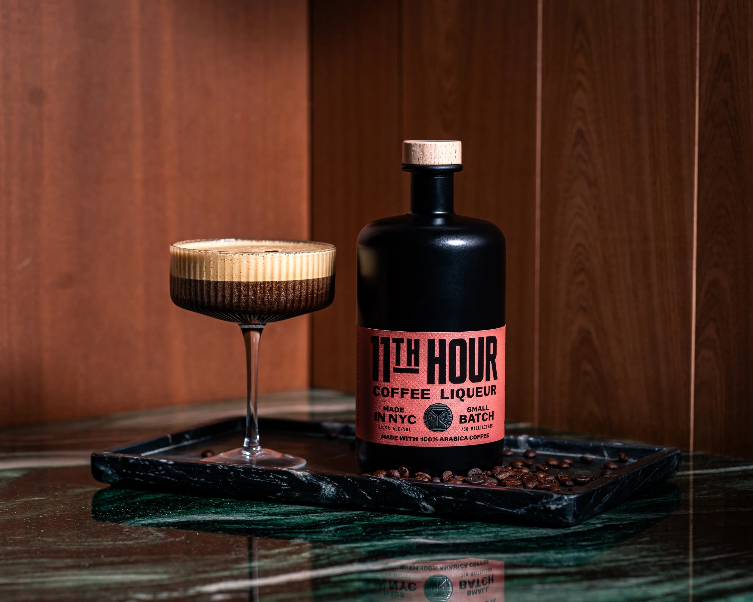 A black bottle labeled "11th Hour Coffee Liqueur" next to a cocktail glass with a coffee drink, on a black marble tray with scattered coffee beans, against a wooden background.