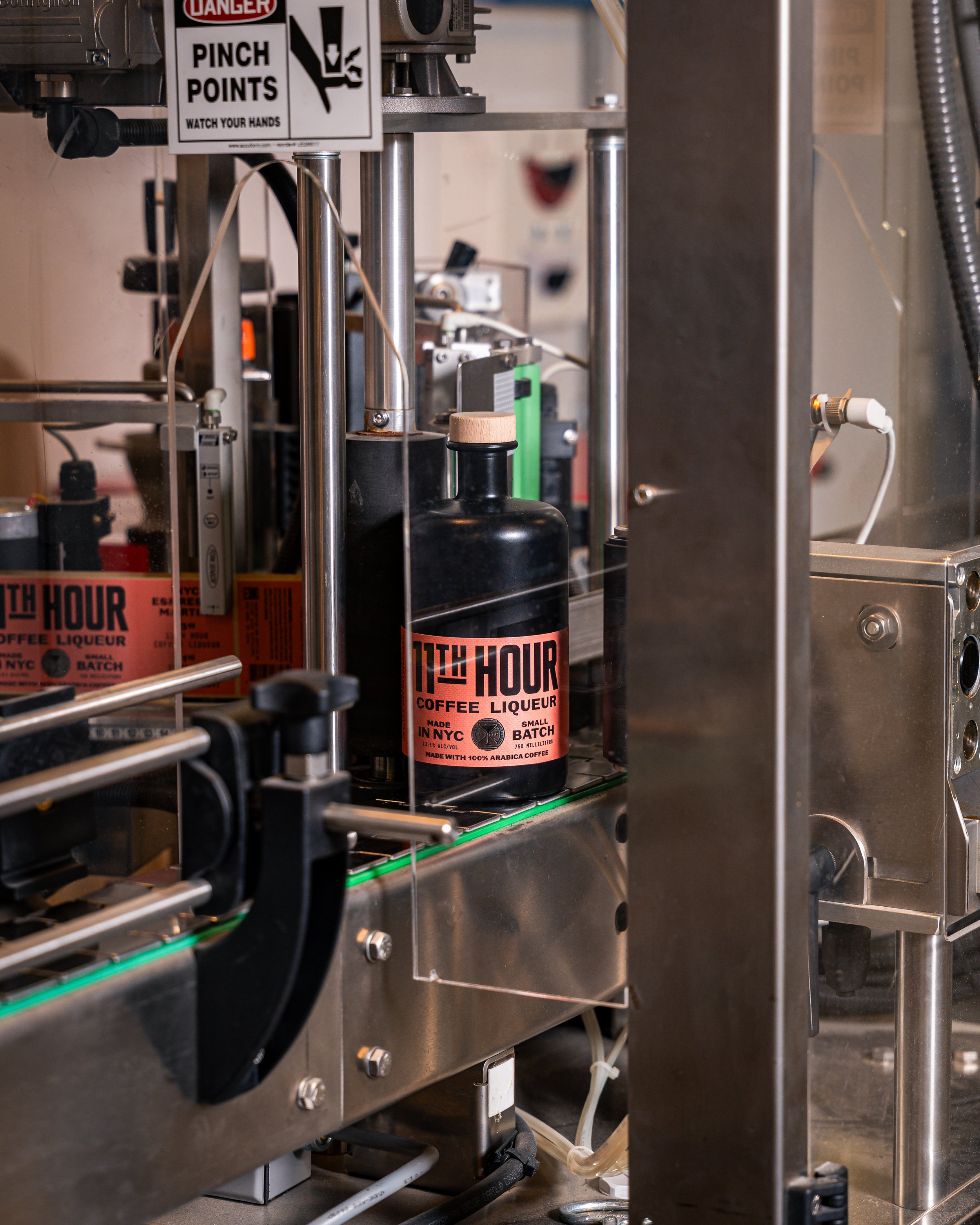 A bottling machine filling a black bottle with a pink label that reads '11th Hour Coffee Liqueur' inside a factory or production facility.