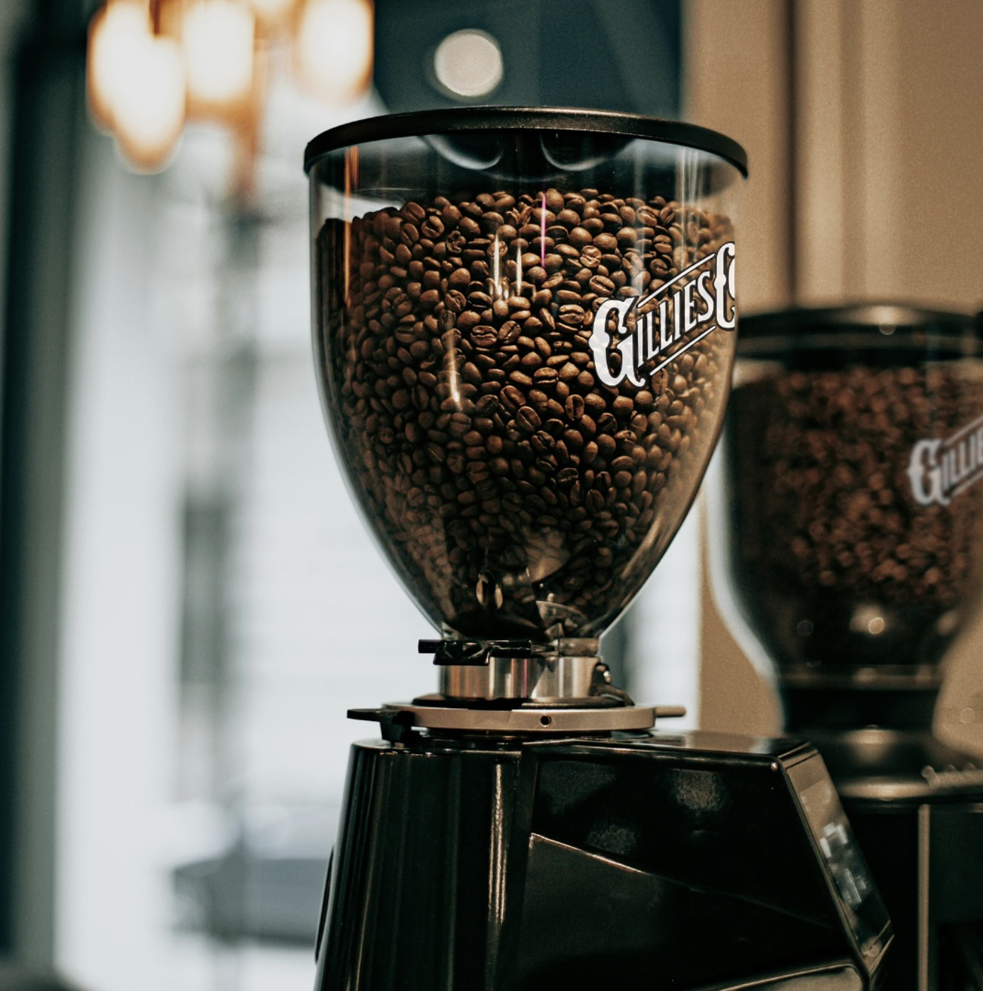 Coffee beans in a clear grinder container with the Glencrest logo, set on a black coffee grinder machine, in a cafe setting.