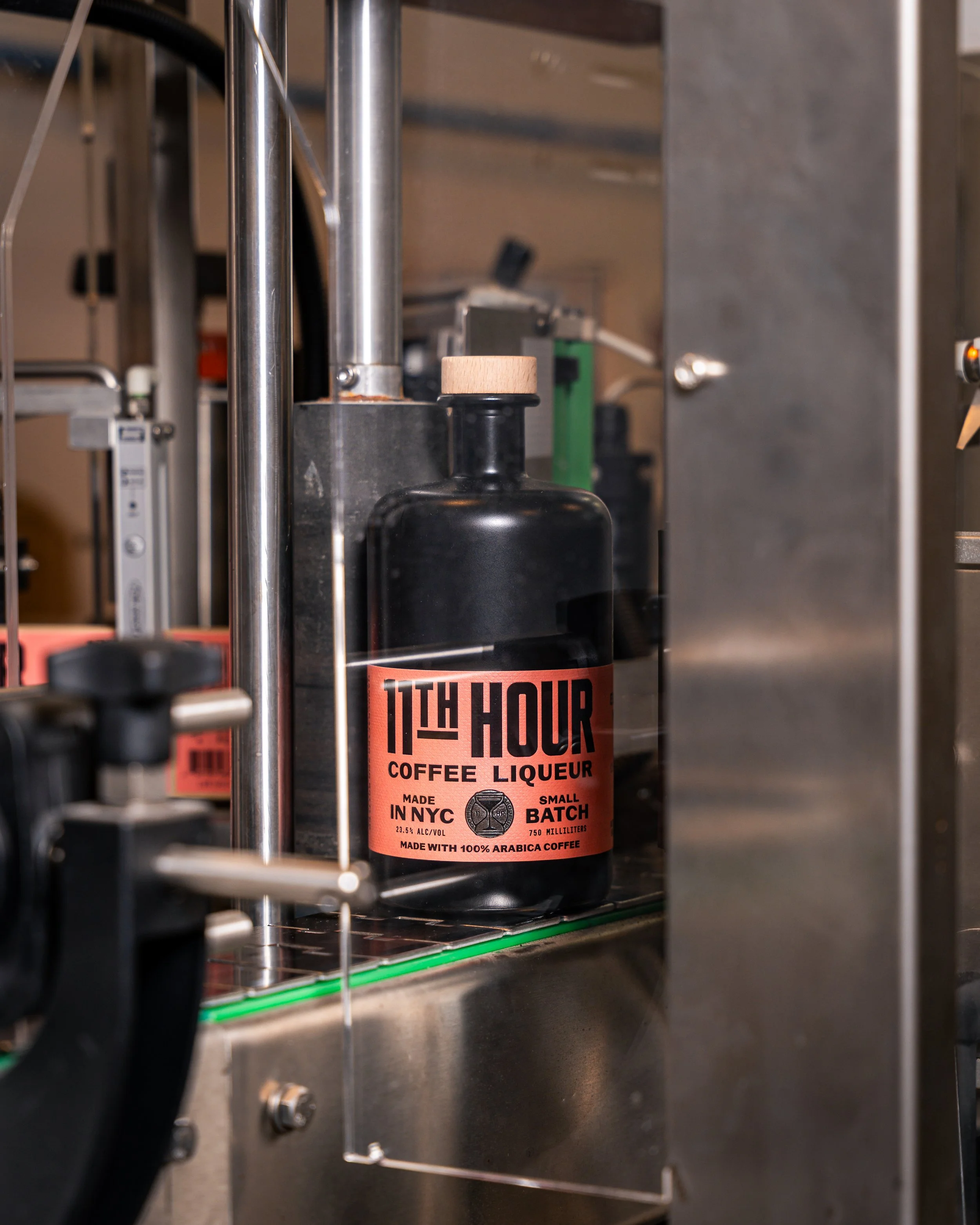 A black bottle labeled '11th Hour Coffee Liqueur' on a stainless steel shelf in a laboratory or industrial setting.