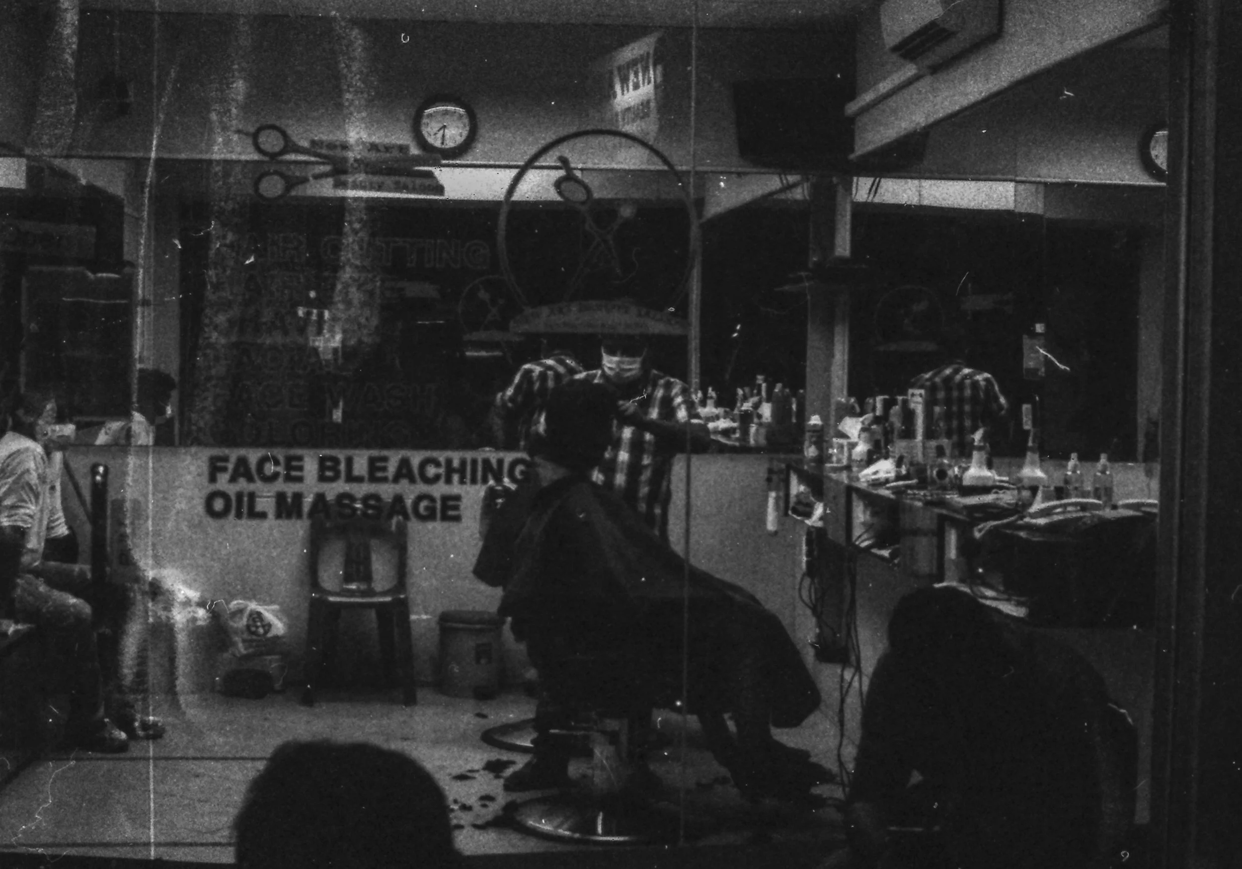 A barber shop with a mirror reflecting a barber cutting a customer's hair, various hair and beauty products on the counter, and a sign that reads "Face Bleaching Oil Massage."