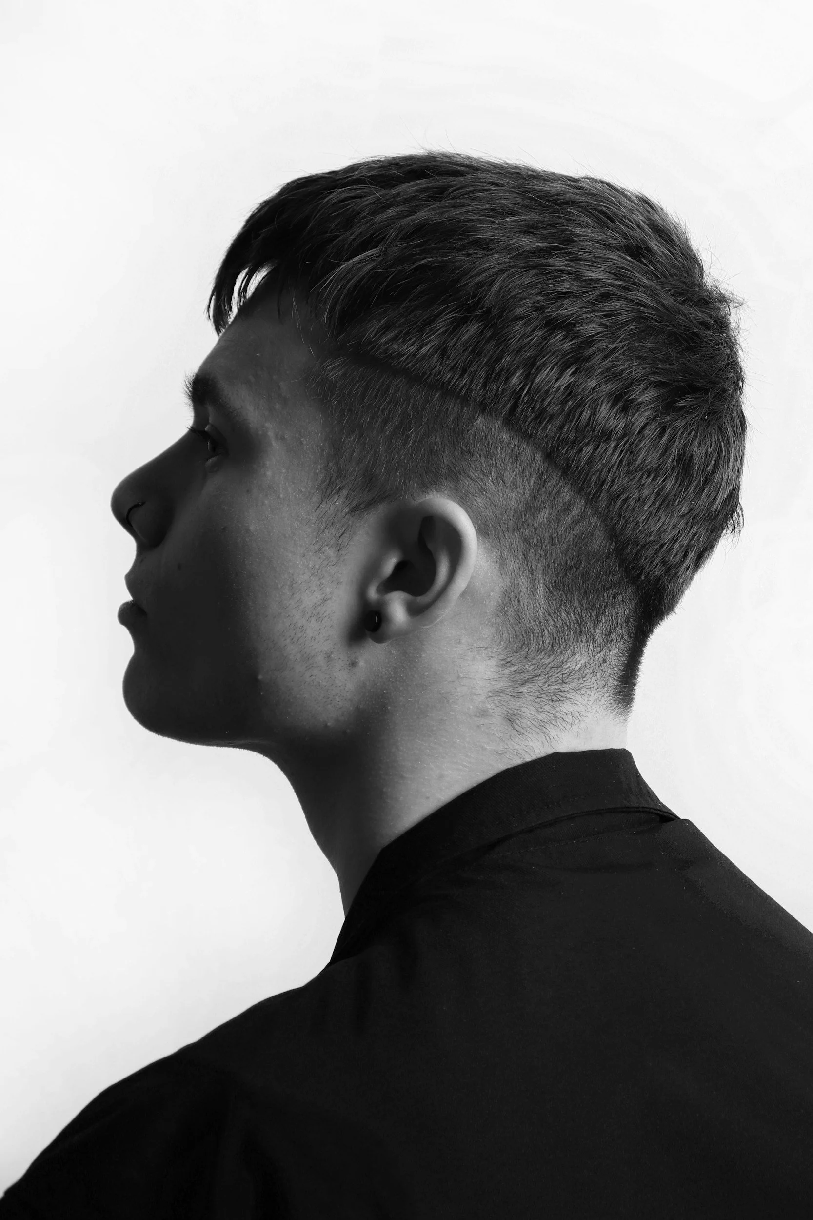Black and white profile of a young man with a modern hairstyle, wearing a dark shirt and small earring, against a plain background.
