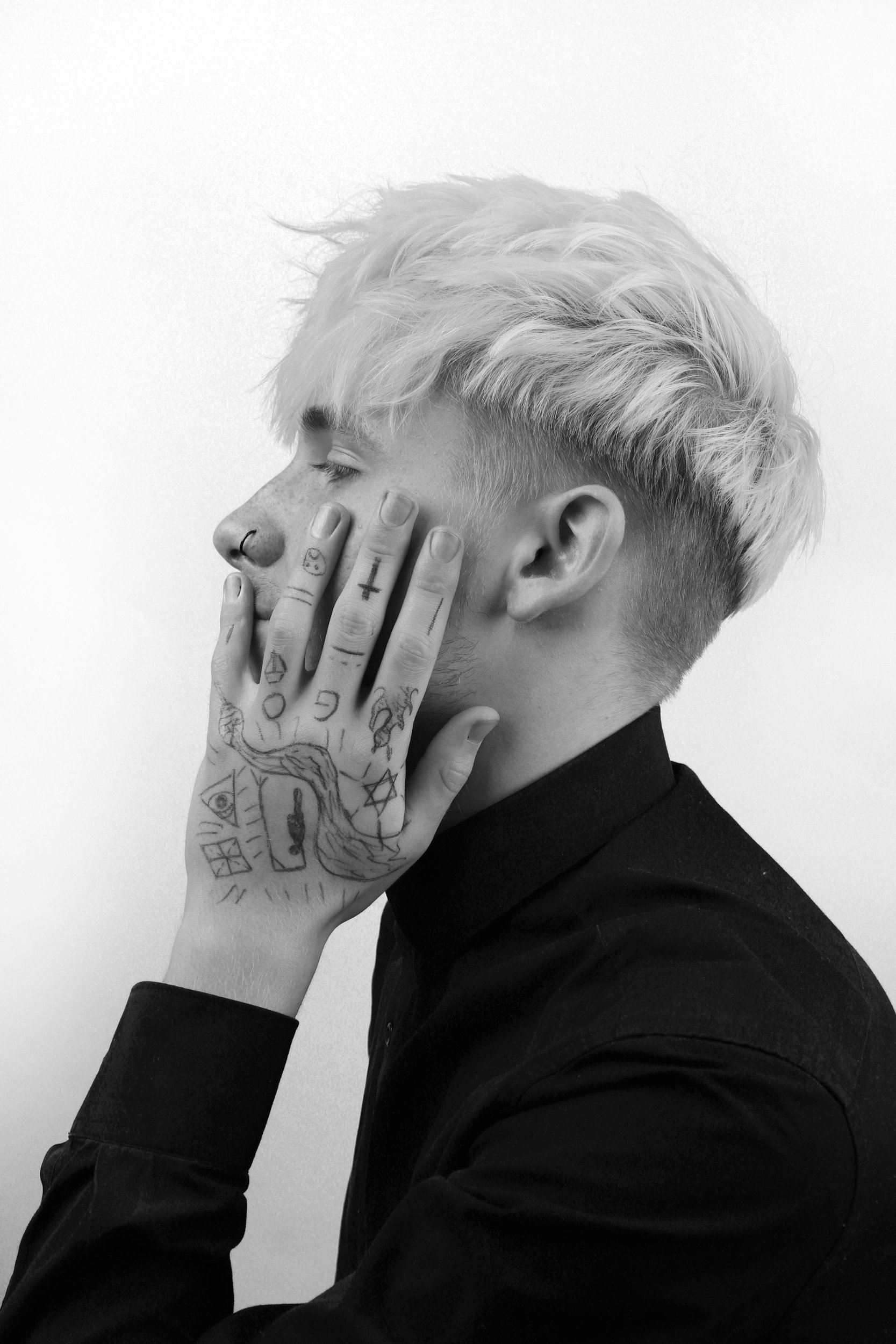 A young person with platinum blonde hair, closed eyes, and multiple tattoos on their hand, wearing a black shirt, resting their chin on their hand against a plain background.