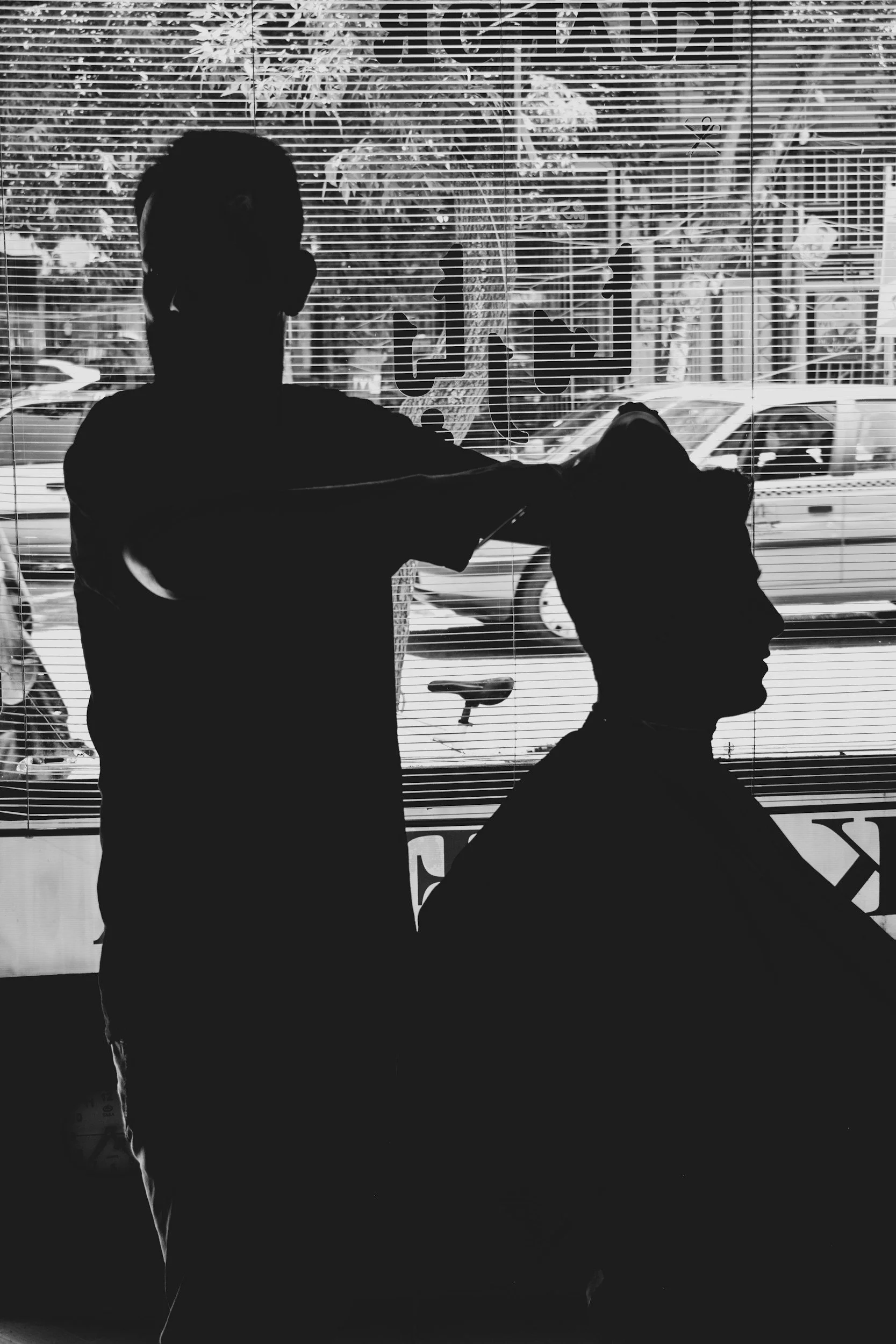 Silhouette of a barber giving a haircut to a customer inside a barbershop, with a window showing parked cars outside.
