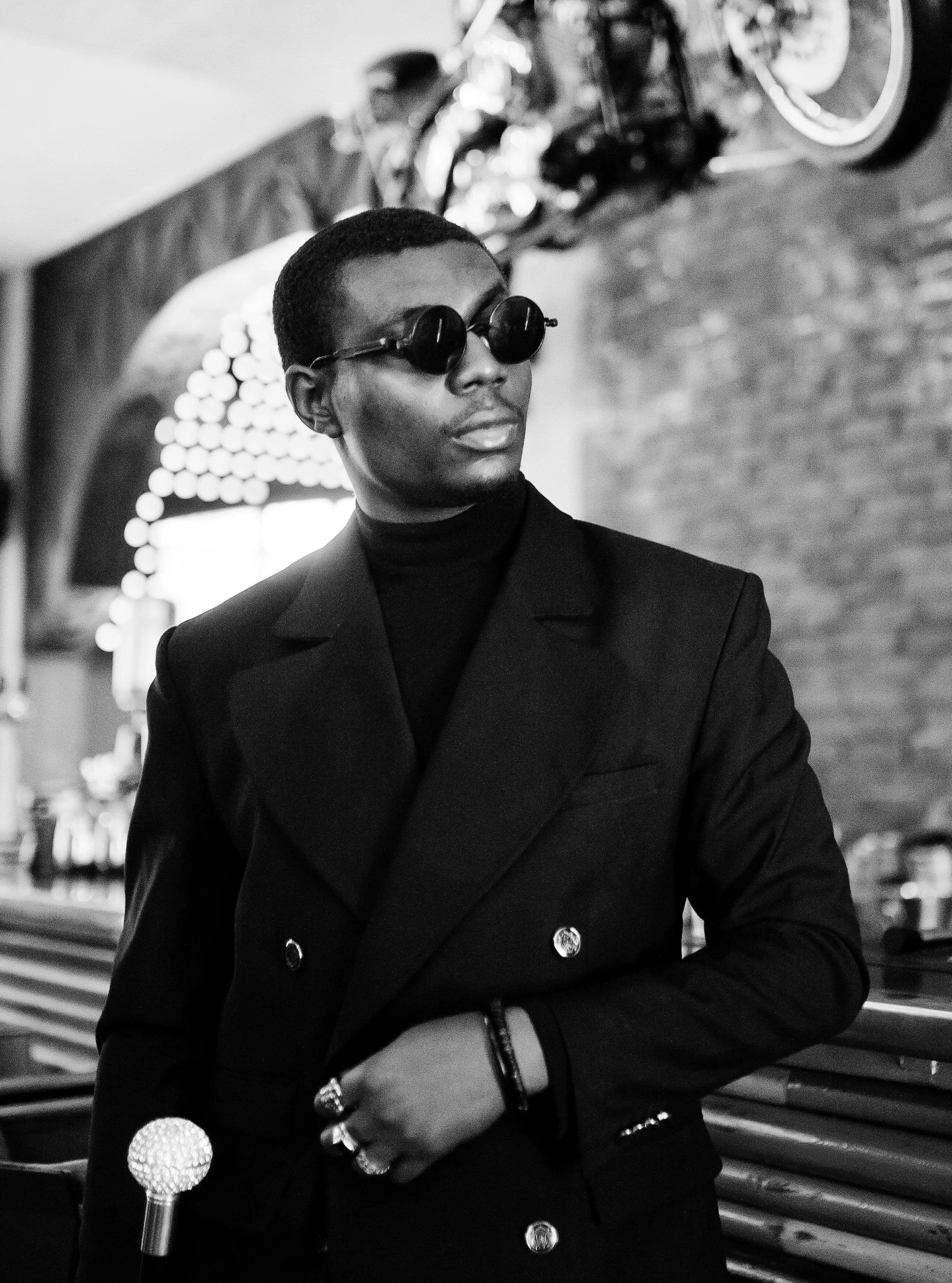 A person in dark clothing and sunglasses standing in an indoor space, with a guitar hanging on the brick wall behind them.