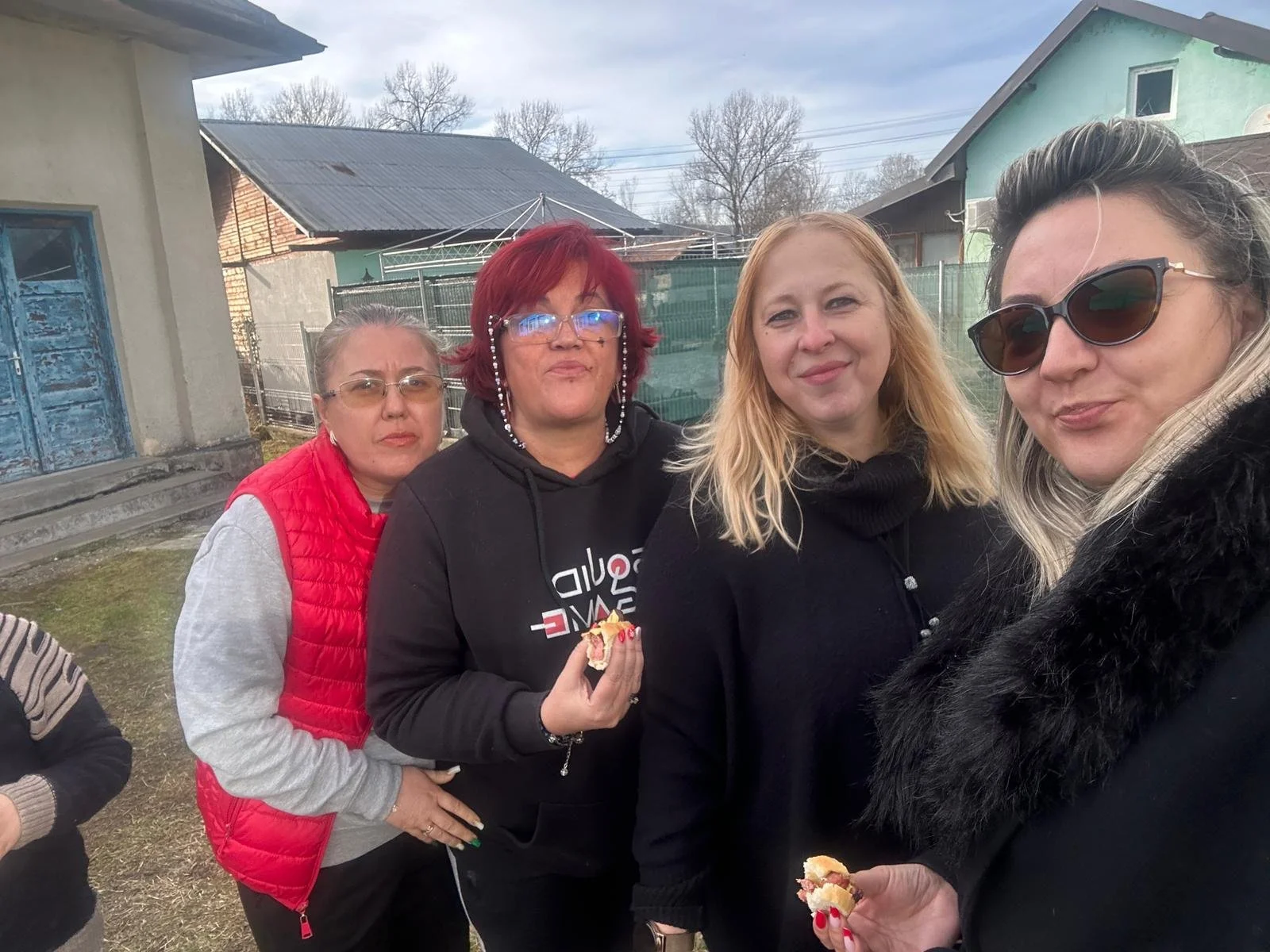 Four women standing outdoors, some holding pieces of cake, in a residential area with houses and a cloudy sky background.