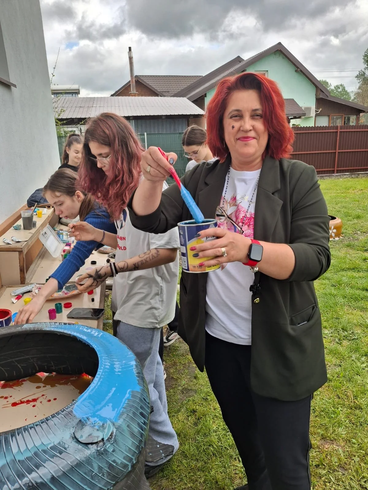 A woman with red hair and a group of children at an outdoor craft activity, with paint supplies and a plastic tub, in a backyard setting.