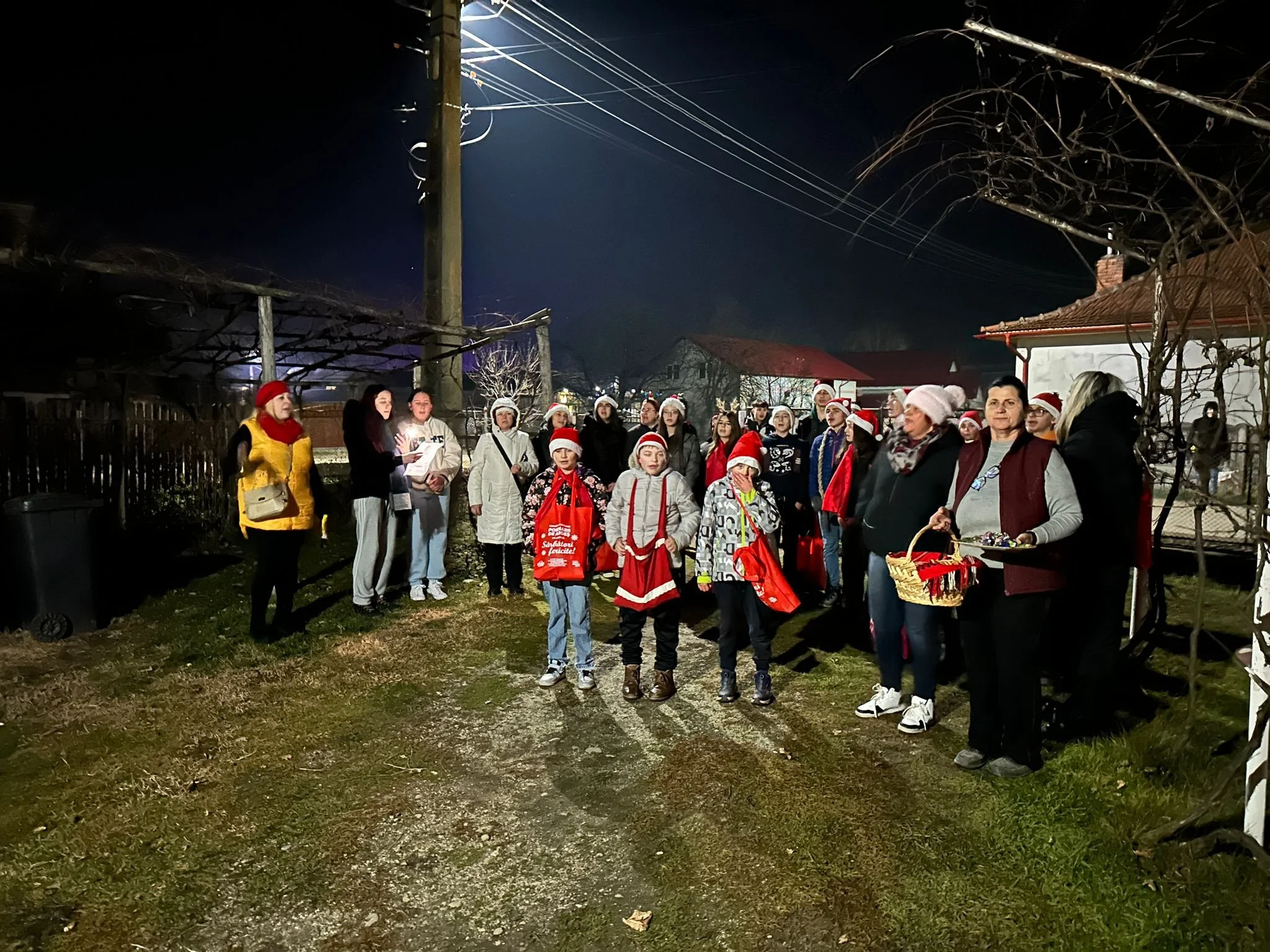 Group of people, including children and adults, wearing Christmas hats and festive clothing, gathered outdoors at night for a holiday celebration.