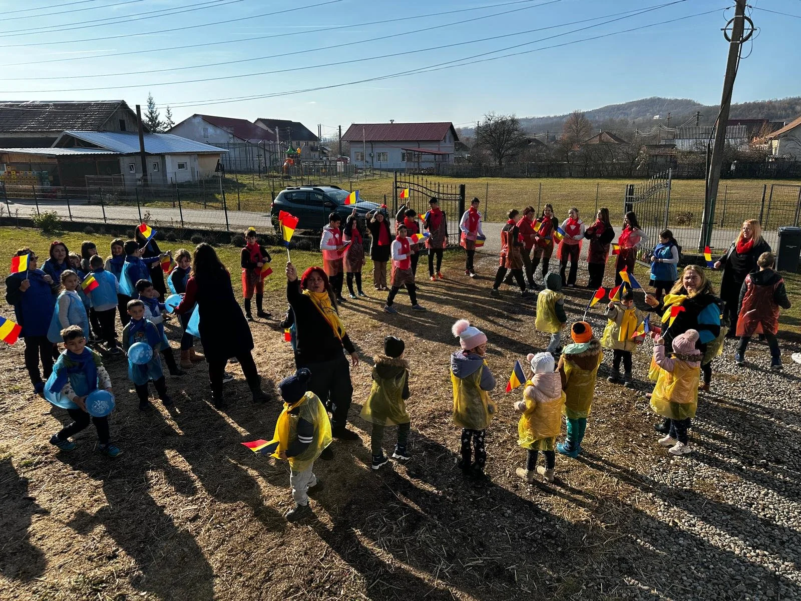 Children and adults outdoors in a yard, some holding Romanian flags, participating in a celebration or event, with houses and a hillside in the background.