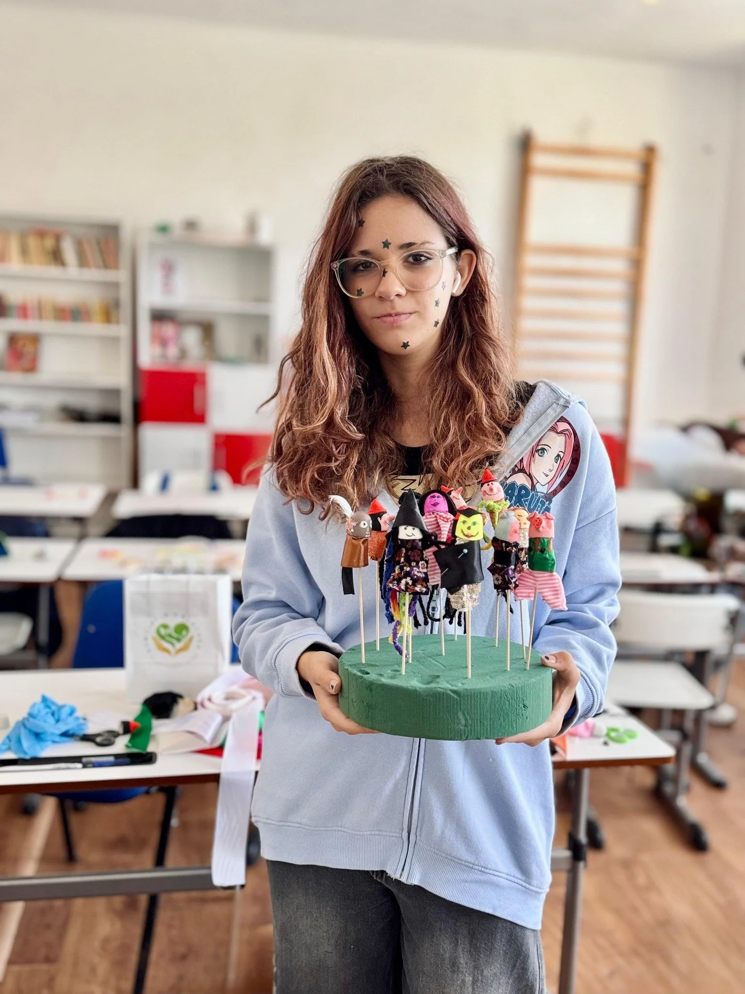 Young woman with glasses and star stickers on her face holding a green cake with Halloween-themed decorations on top.