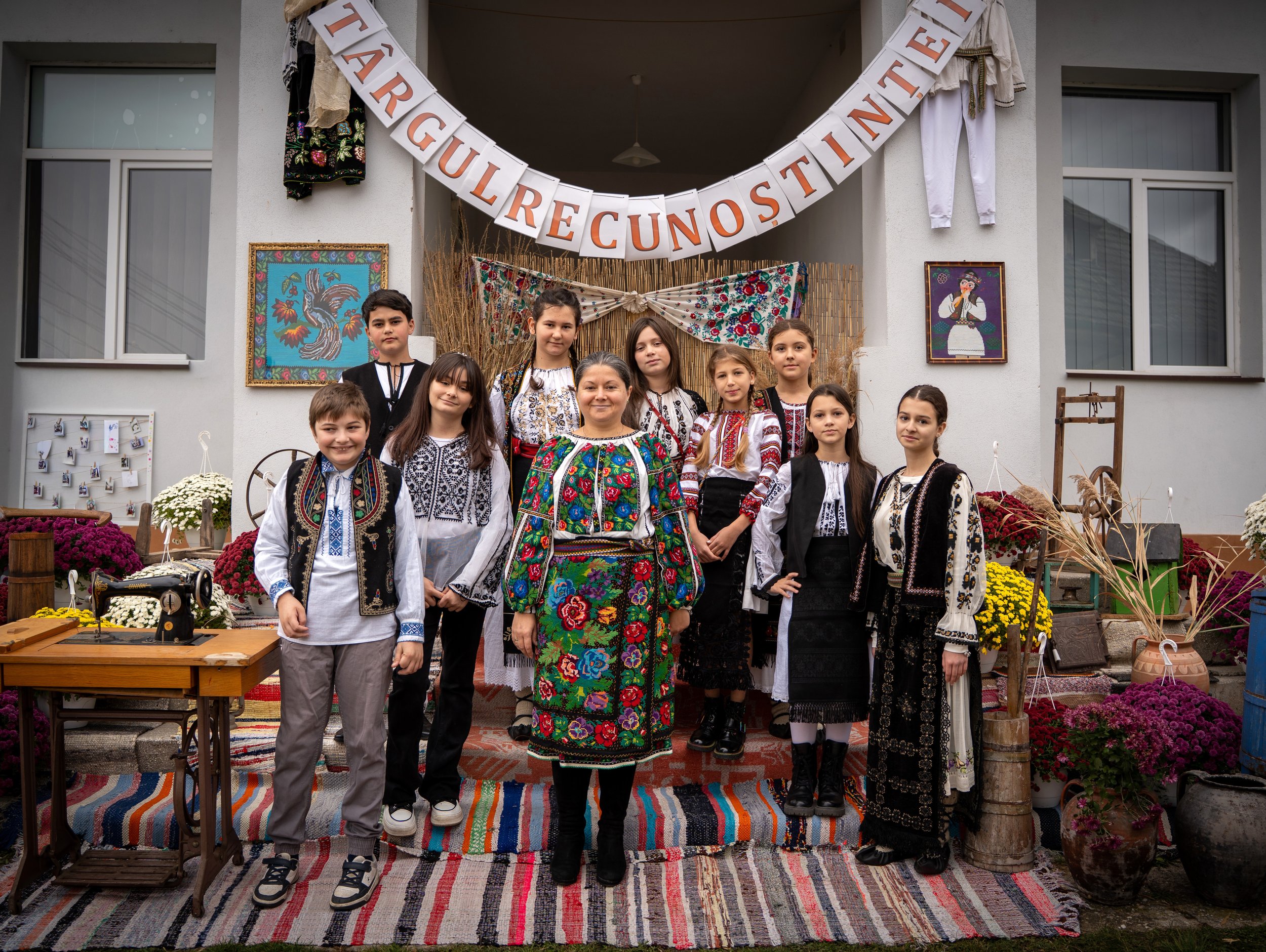 Group of children and a woman dressed in traditional Romanian clothing, standing on a colorful woven rug outdoors in front of a decorated wall with a banner that reads 'ŢARIGUL RE CUNOSTINŢE TE'. The scene includes traditional crafts and decorations,