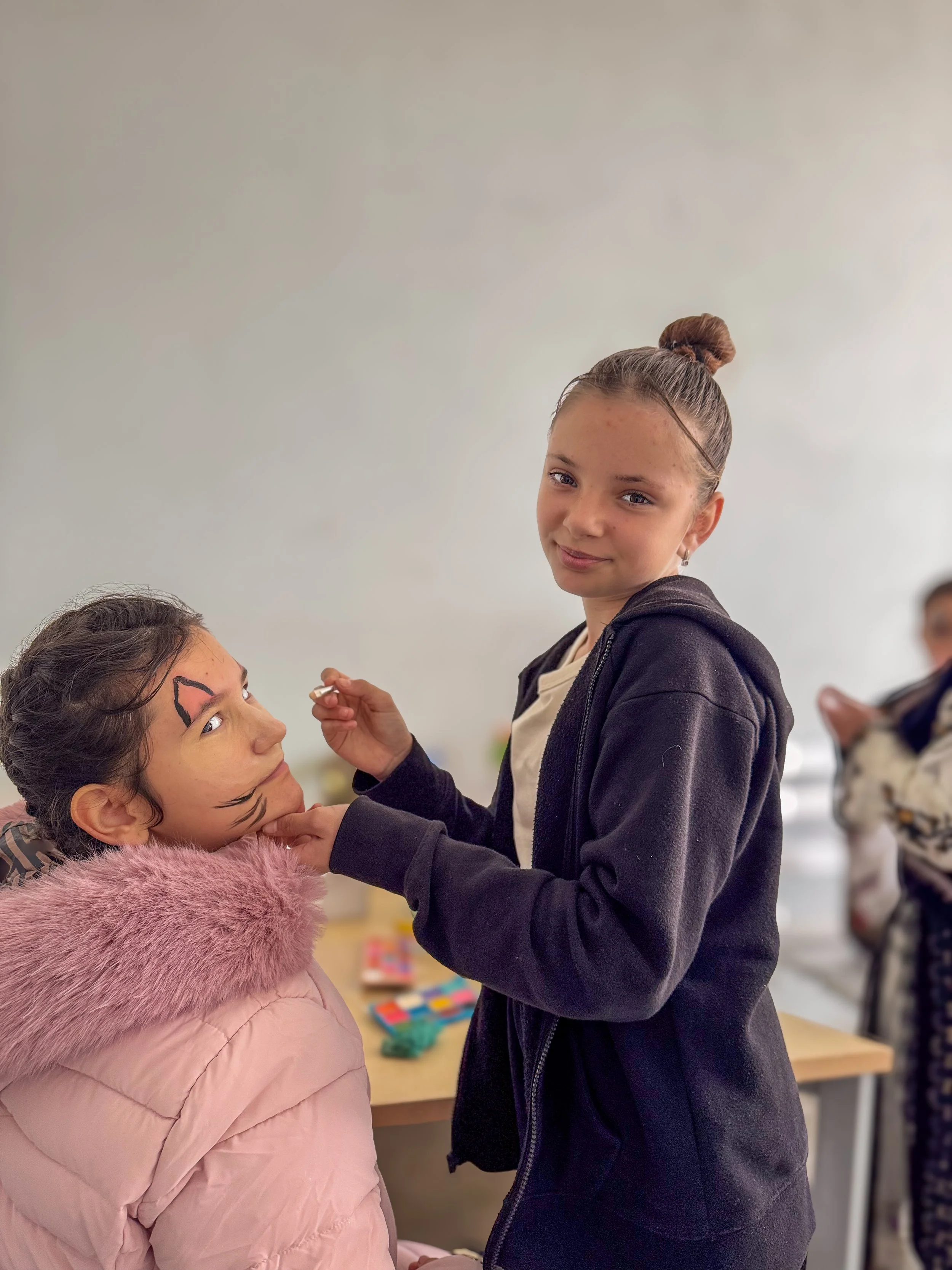 A girl with face paint, dressed in a pink coat with a furry collar, looks up at a standing girl with a bun hairstyle, wearing a black hoodie, as she paints a face picture on her face. There is a blurred person in the background.