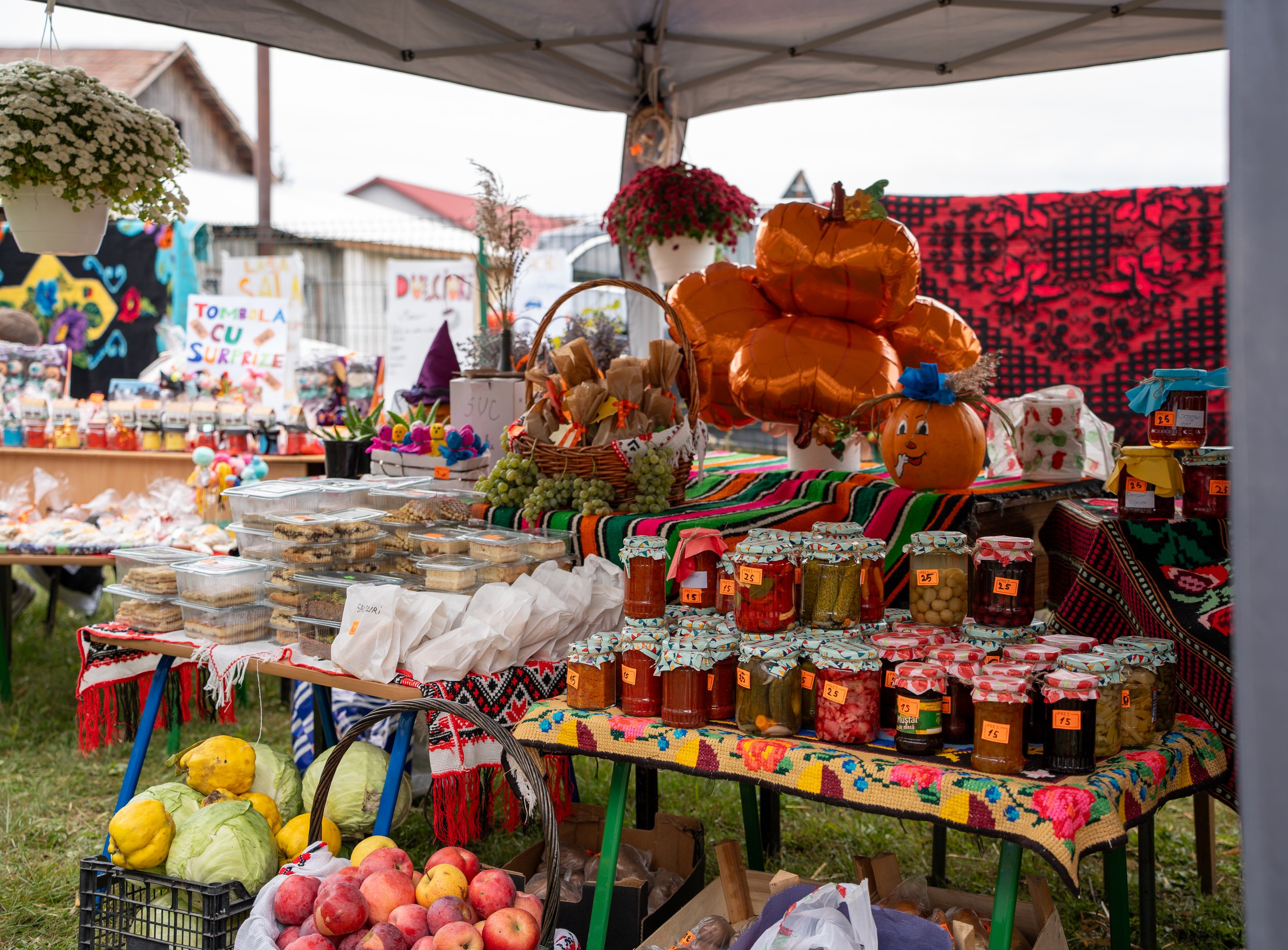 Outdoor marketplace stall selling jars of preserves and jams, pumpkins, apples, and decorative balloon pumpkins.