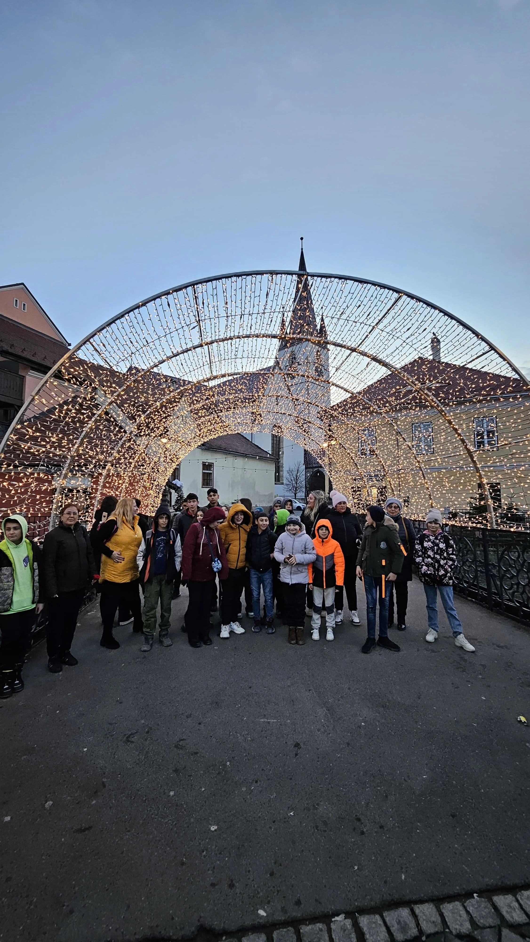 Group of people standing under a festive archway decorated with string lights during evening.