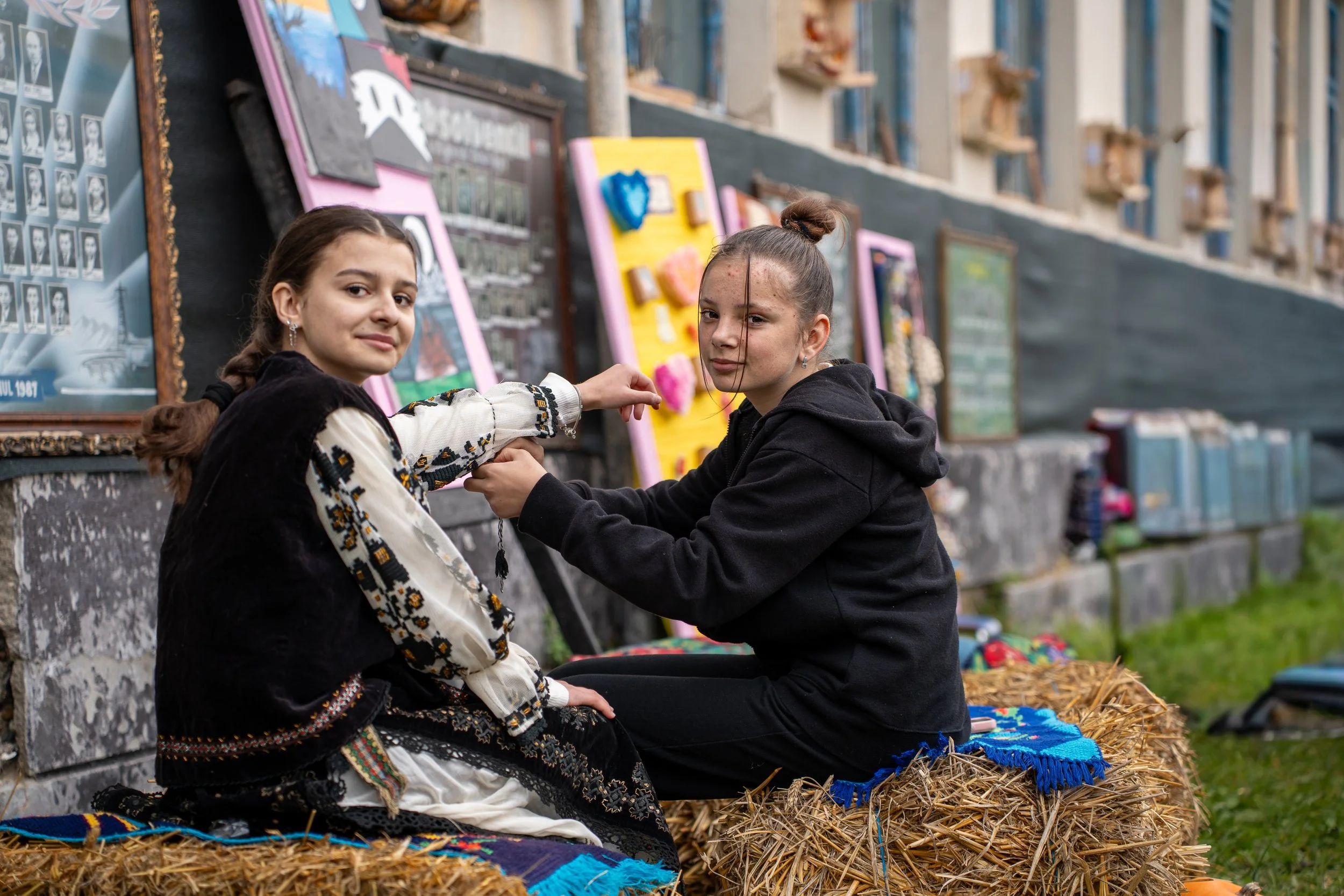 Two girls sitting on hay bales in front of a wall of framed photos and artwork, exchanging necklaces outdoors.