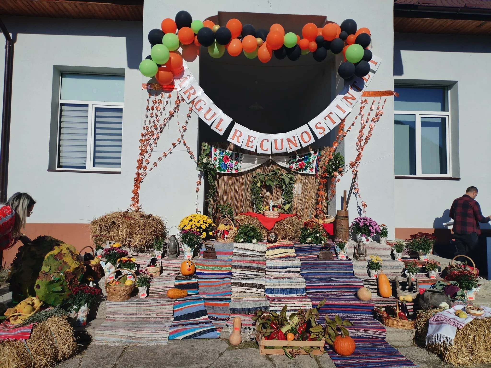 Festive outdoor display with hay bales, pumpkins, gourds, flowers, and decorative textiles on a porch, with a banner that reads "Trigual Recunostin" and colorful balloons overhead, celebrating autumn or a harvest festival.