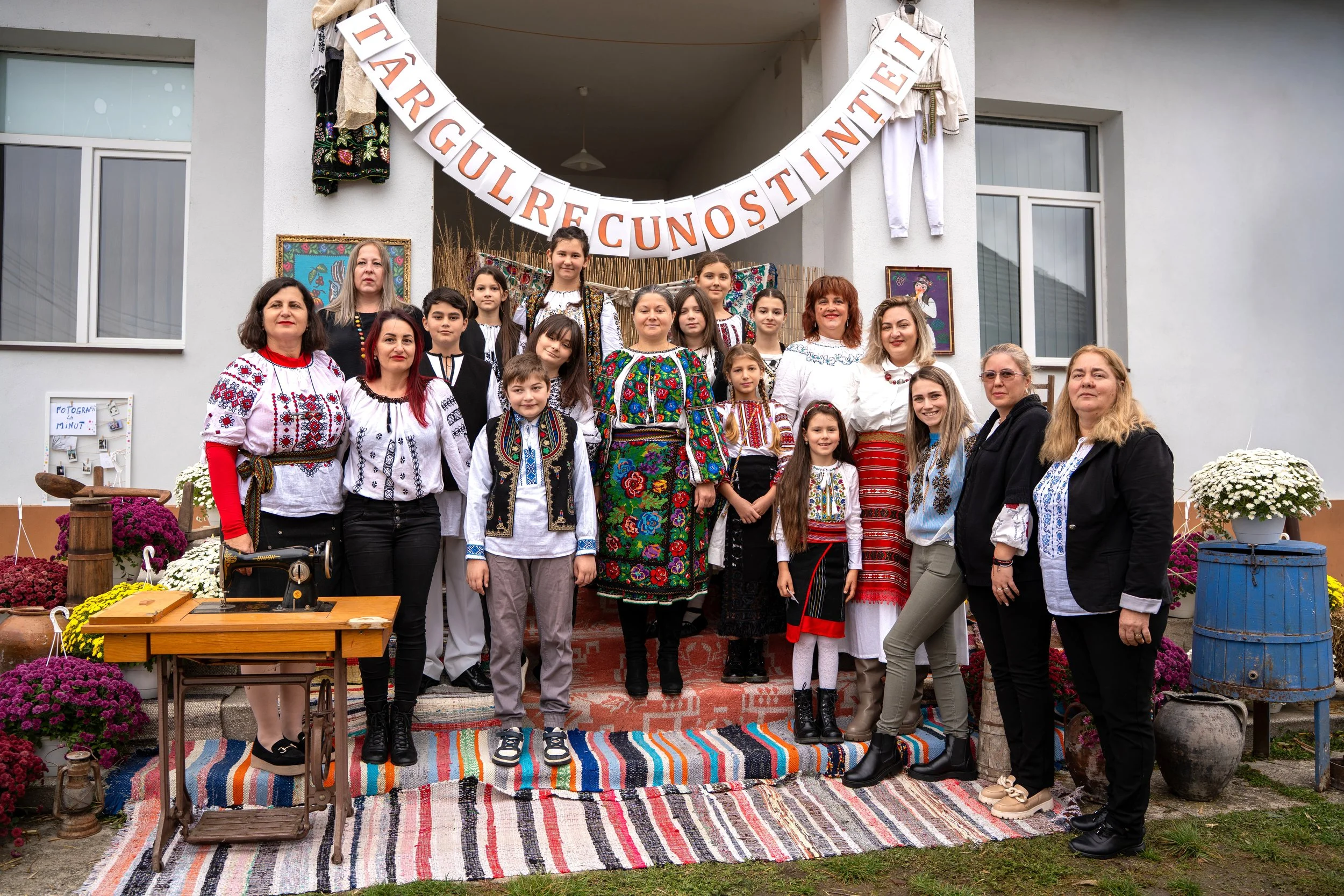 Group of children and adults dressed in traditional embroidered clothing, standing on a decorated porch with colorful rugs and flowers, celebrating a cultural event with a banner above that reads 'TÂRGUL RECU NOS TINTEI'.