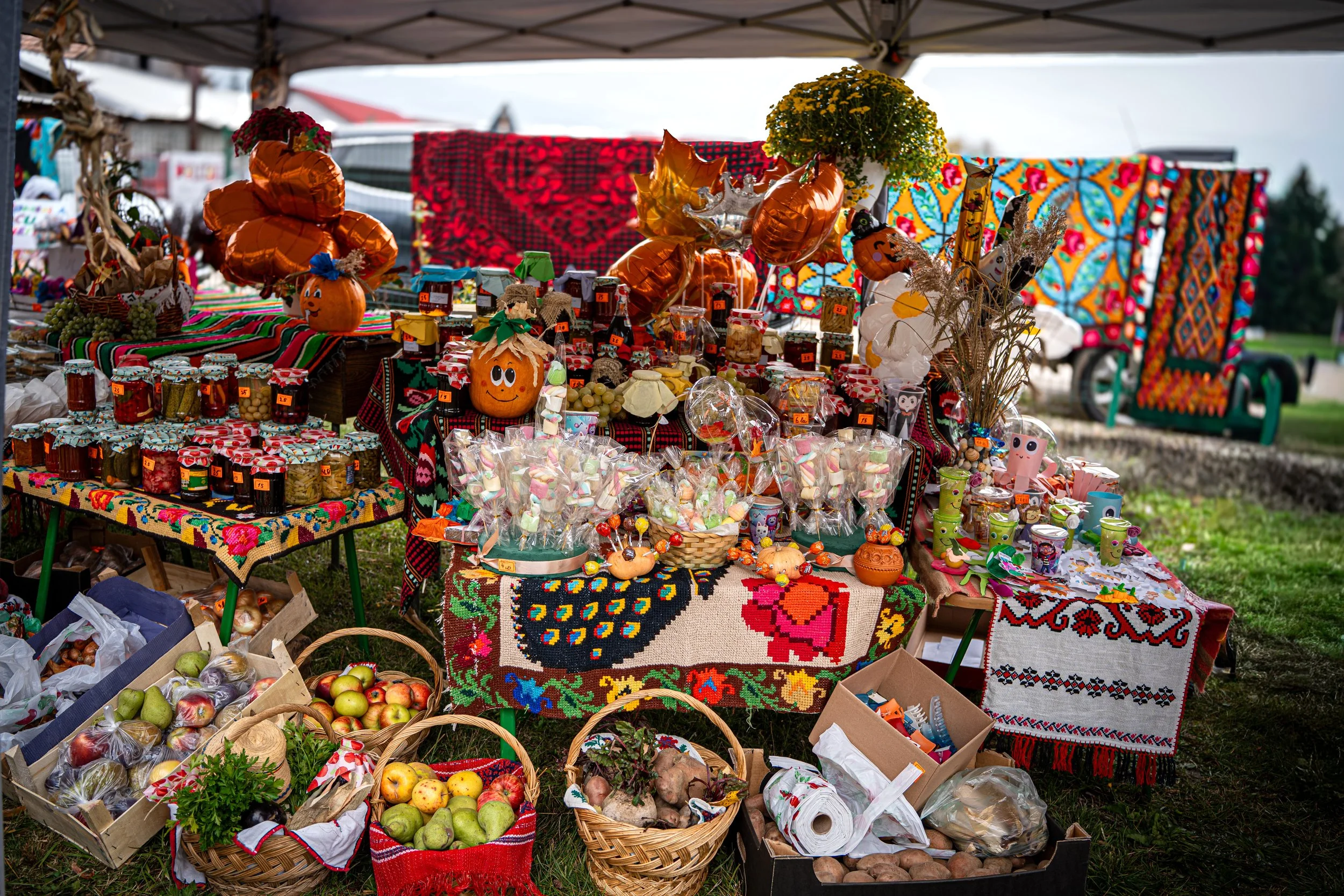 An outdoor market stall displaying colorful homemade preserves, baked goods, and seasonal decorations, with woven tablecloths and pumpkin-themed items.