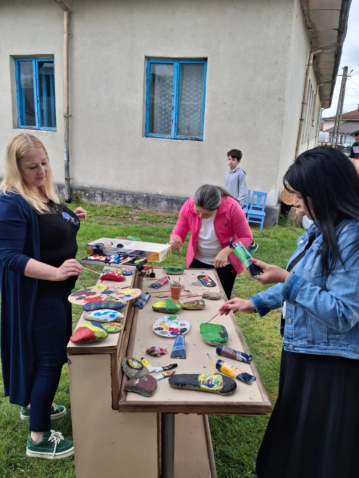 People painting decorated rocks on a table outdoors near a building.