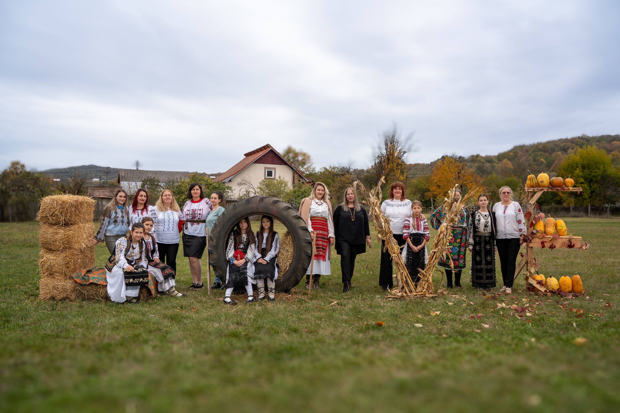 Group of people in traditional clothing gathering outdoors during autumn, with hay bales, pumpkins, and autumn decorations.