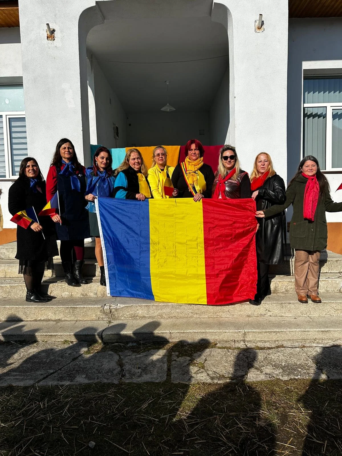 Group of women standing on steps holding a large Romanian flag and small Romanian flags outdoors in front of a white building.