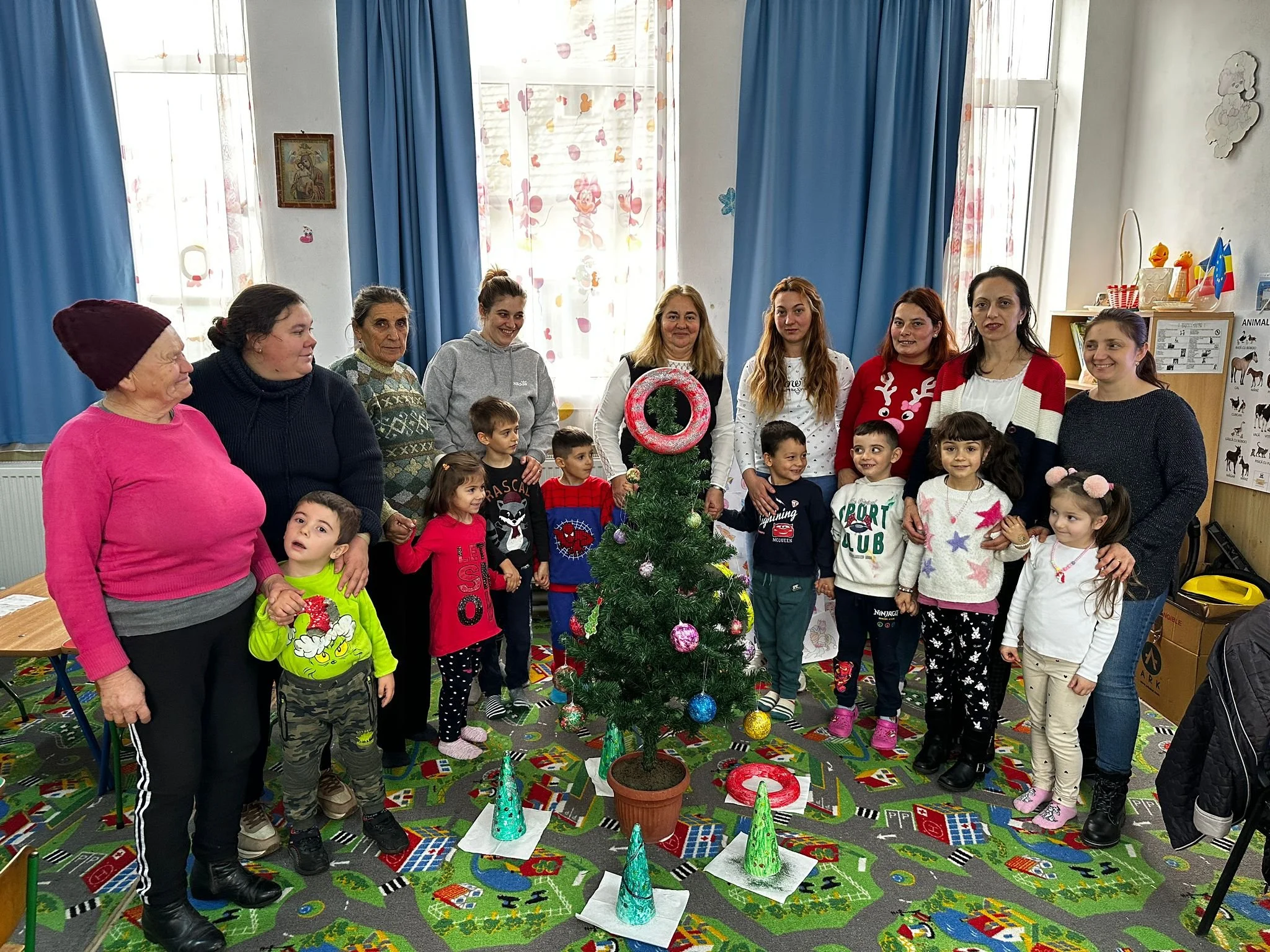 A group of children and adults standing around a decorated small Christmas tree in a room with colorful curtains and a patterned carpet.