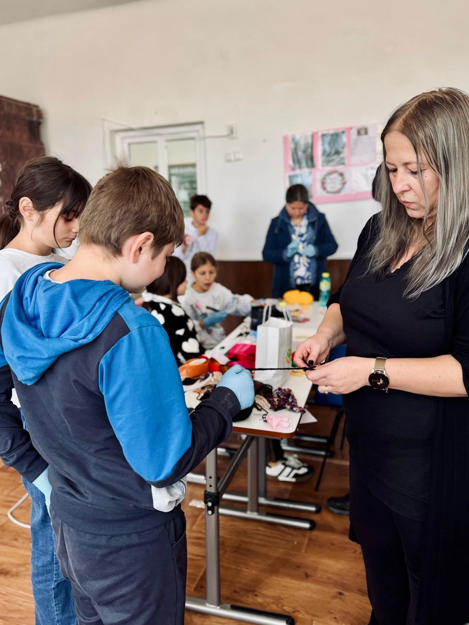 A group of children and an adult woman in a classroom or community center, with tables and crafts supplies, engaging in an activity.