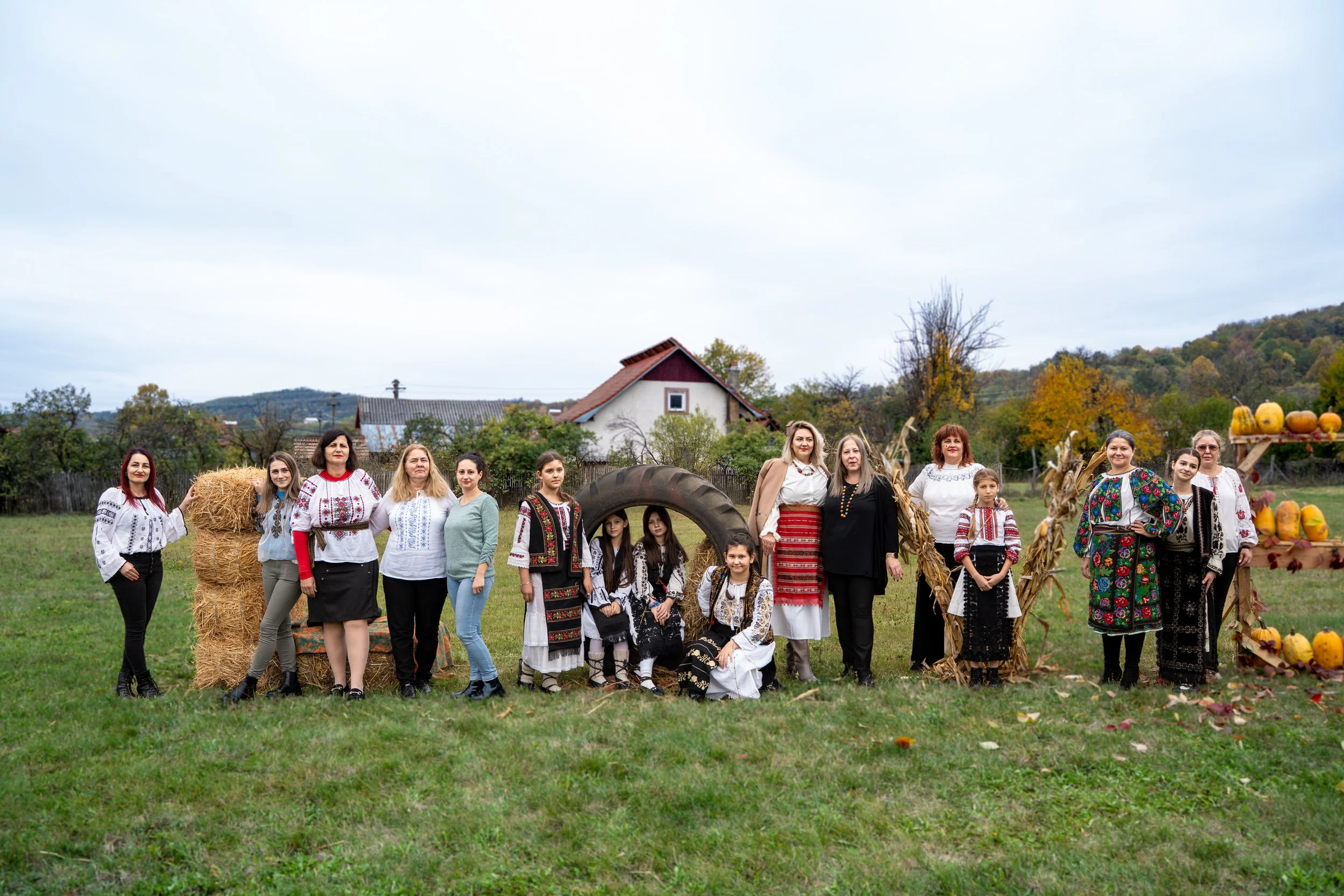 A group of people dressed in traditional Eastern European or Ukrainian clothing, standing outdoors on a grassy field with autumn scenery, hay bales, and pumpkins in the background, possibly during a harvest celebration.