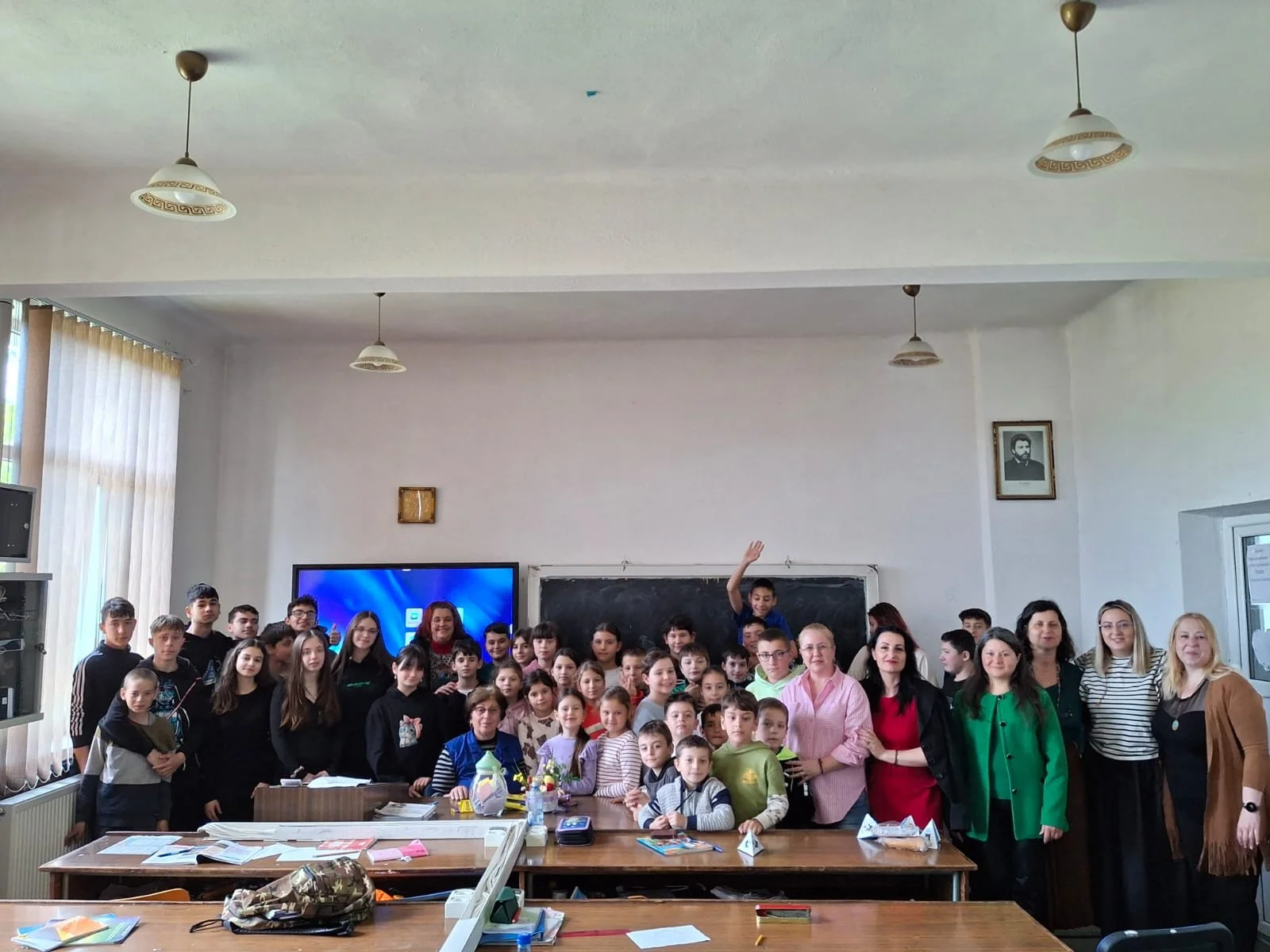 A large group of children and adults gathered in a classroom, posing for a photo. There are tables with books and supplies in the foreground, a blackboard behind them, and a TV screen displaying a blue background. The classroom has white walls, windo