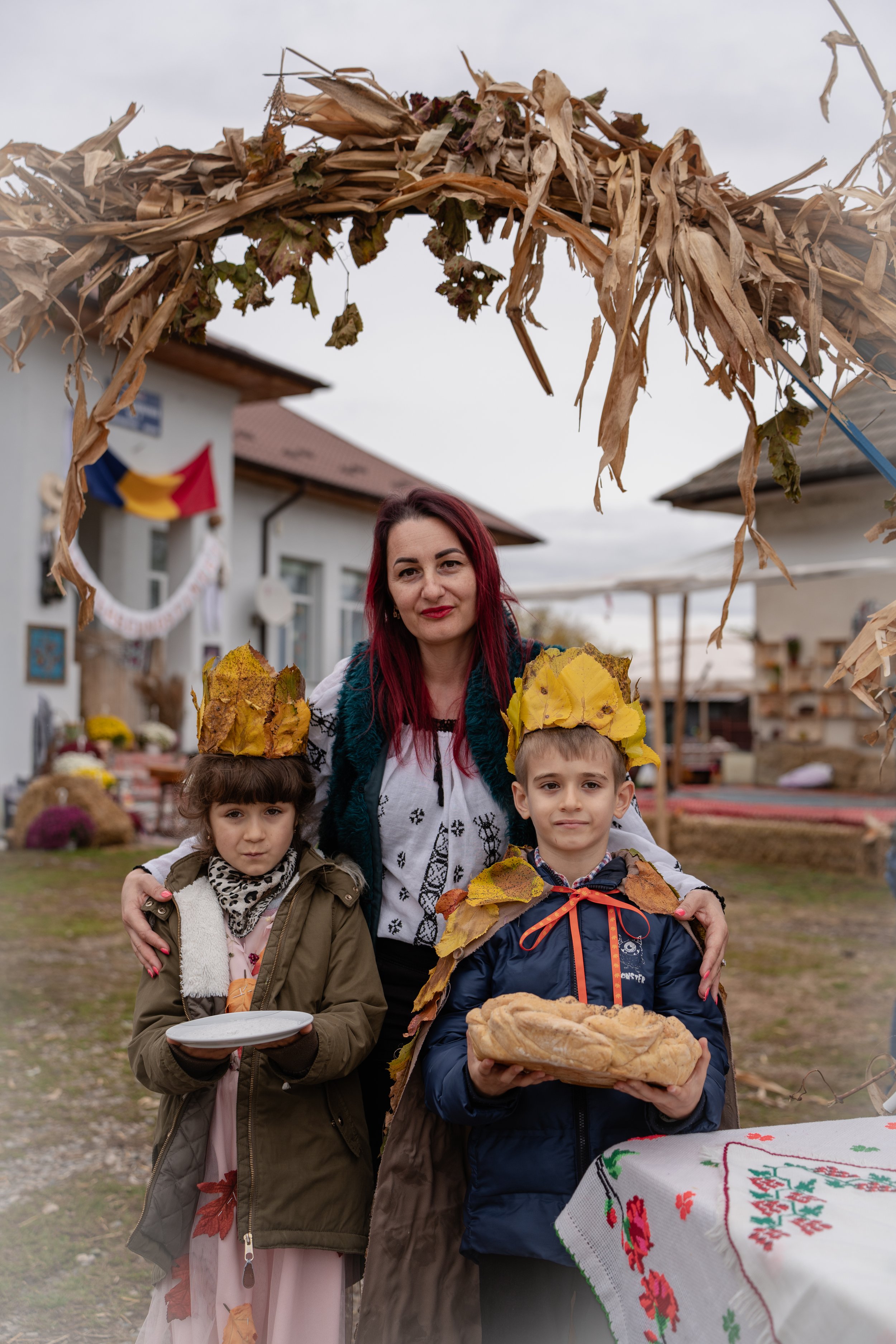 A woman and two children pose outdoors under a decorative arch made of dried corn stalks and autumn leaves. The woman has red hair and is smiling, with her arms around the children. The girl on the left holds a plate, and the boy on the right holds a